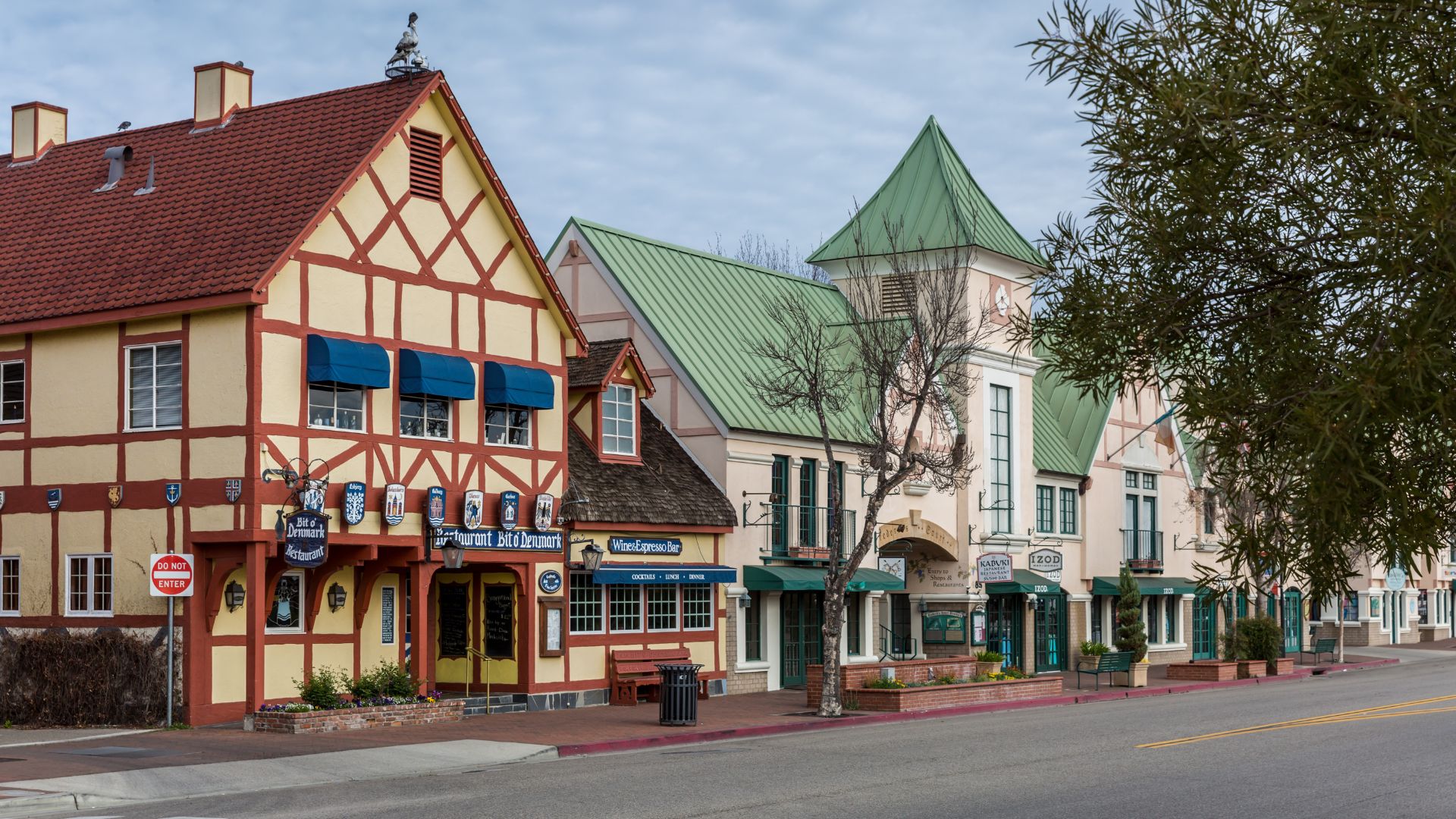 A street view of Solvang, California, featuring colorful buildings with Danish-style architecture, including half-timbered facades and steep gabled roofs, under a clear sky. The buildings house businesses like a restaurant and a wine & espresso bar, and the scene includes a street with markings and a tree.