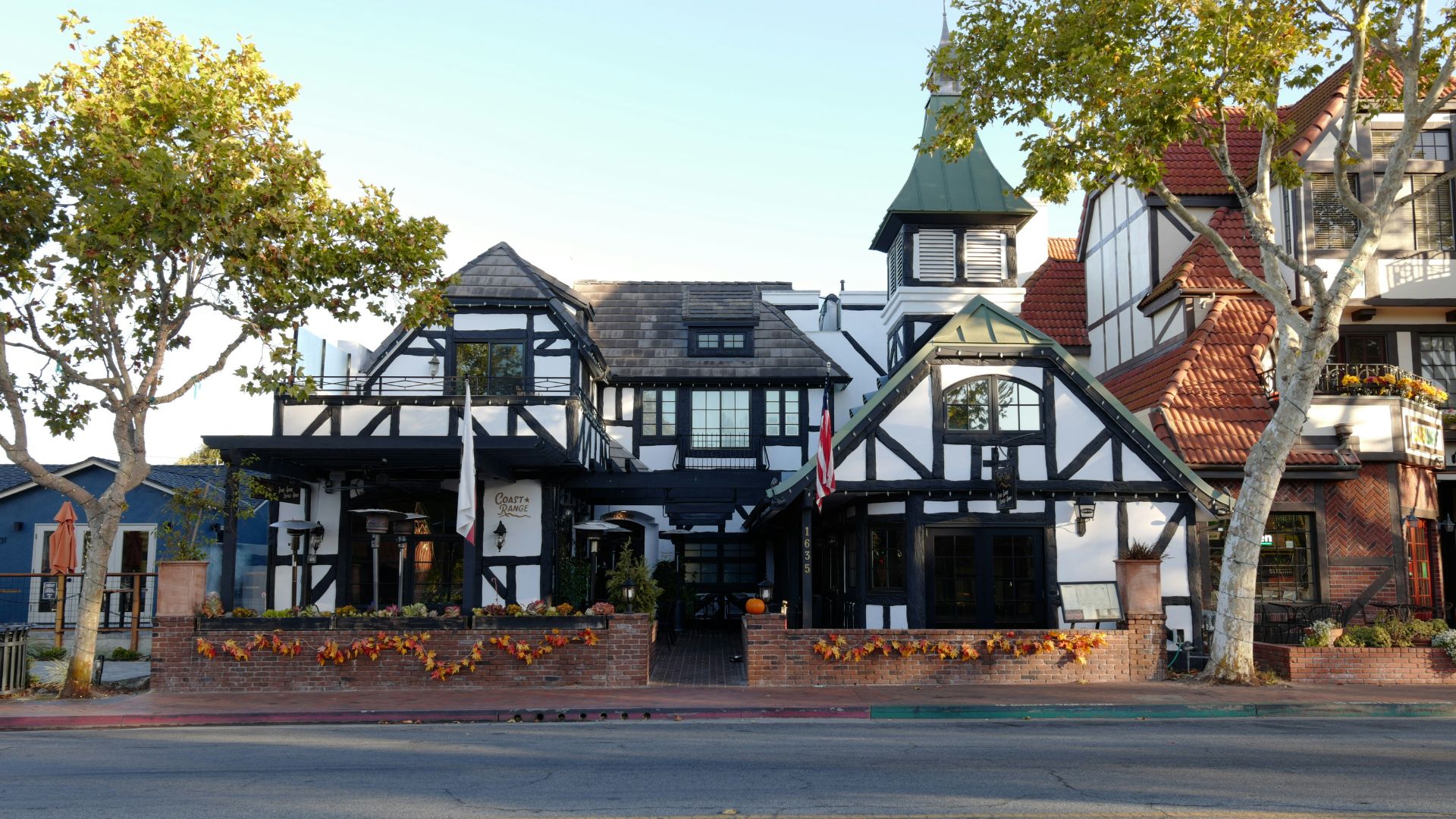 A daytime shot of a half-timbered building with a gabled roof and a tower, decorated with pumpkins along a brick wall, situated on a street in Solvang, California.