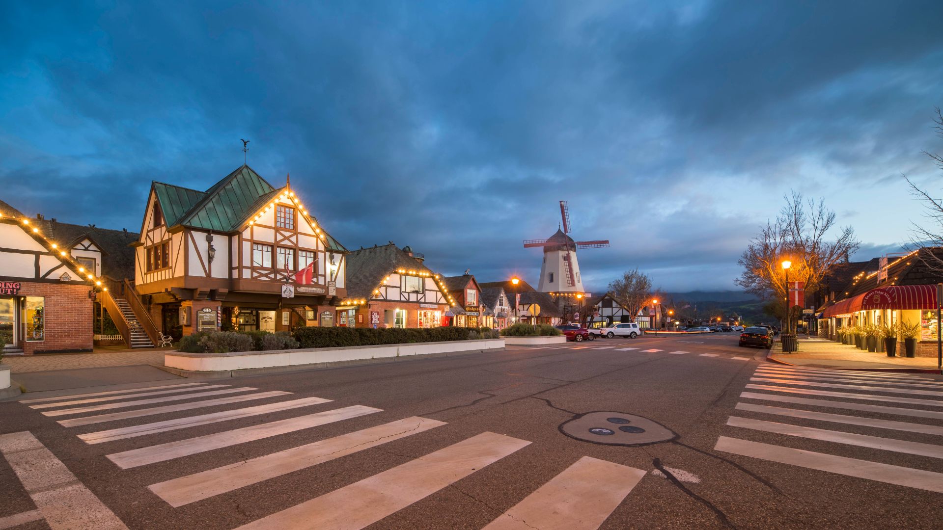 A nighttime view of a quaint street in Solvang, California, featuring buildings with Danish architectural elements, illuminated storefronts, a prominent windmill in the background, and crosswalks in the foreground, all under a cloudy sky. 