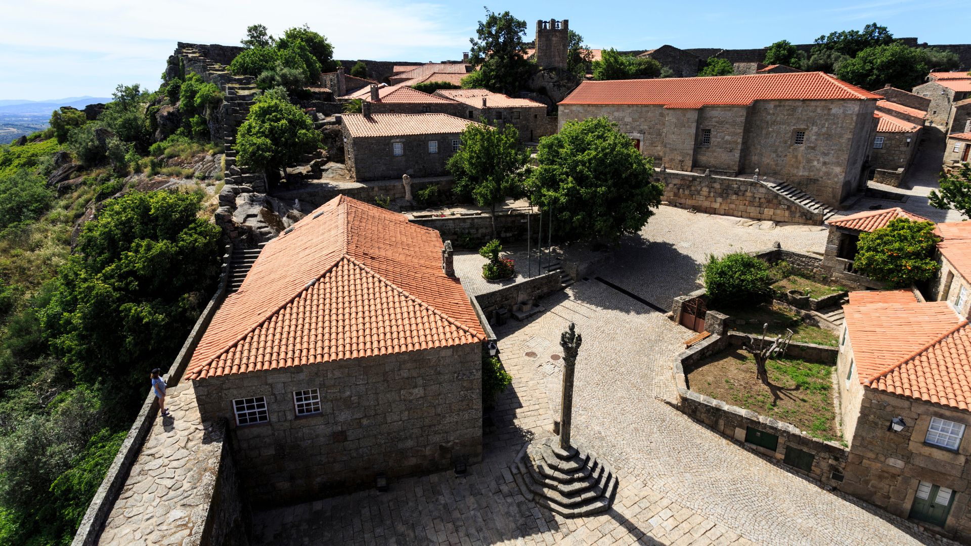 A high-angle view of Sortelha, a historic Portuguese village, showcasing its traditional stone buildings with red-tiled roofs, a central paved square with a stone pillory, and surrounding green trees and rocky terrain, all under a clear sky.
