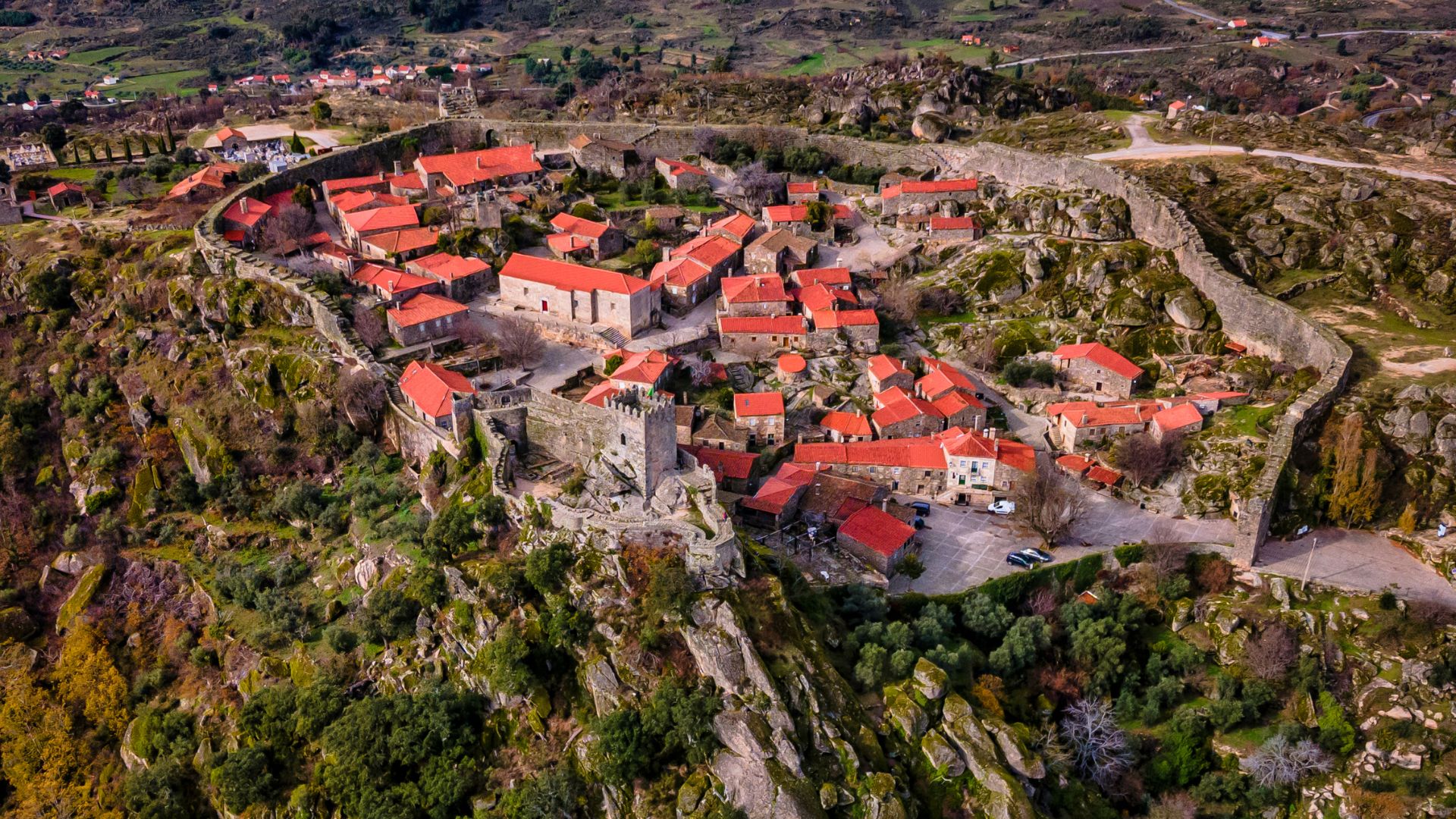 An aerial view of Sortelha, a historic walled village in Portugal, showcasing its medieval architecture with red-tiled roofs nestled within a stone wall on a rocky, tree-covered landscape.