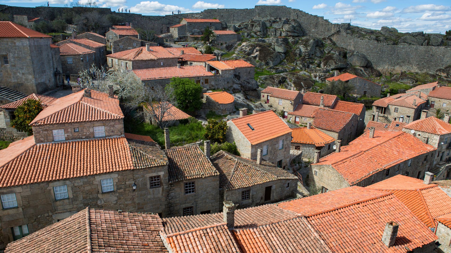 An aerial view of Sortelha, a historic medieval village in Portugal, featuring stone houses with orange-tiled roofs nestled within a defensive wall and overlooked by a castle on a rocky outcrop under a blue sky.