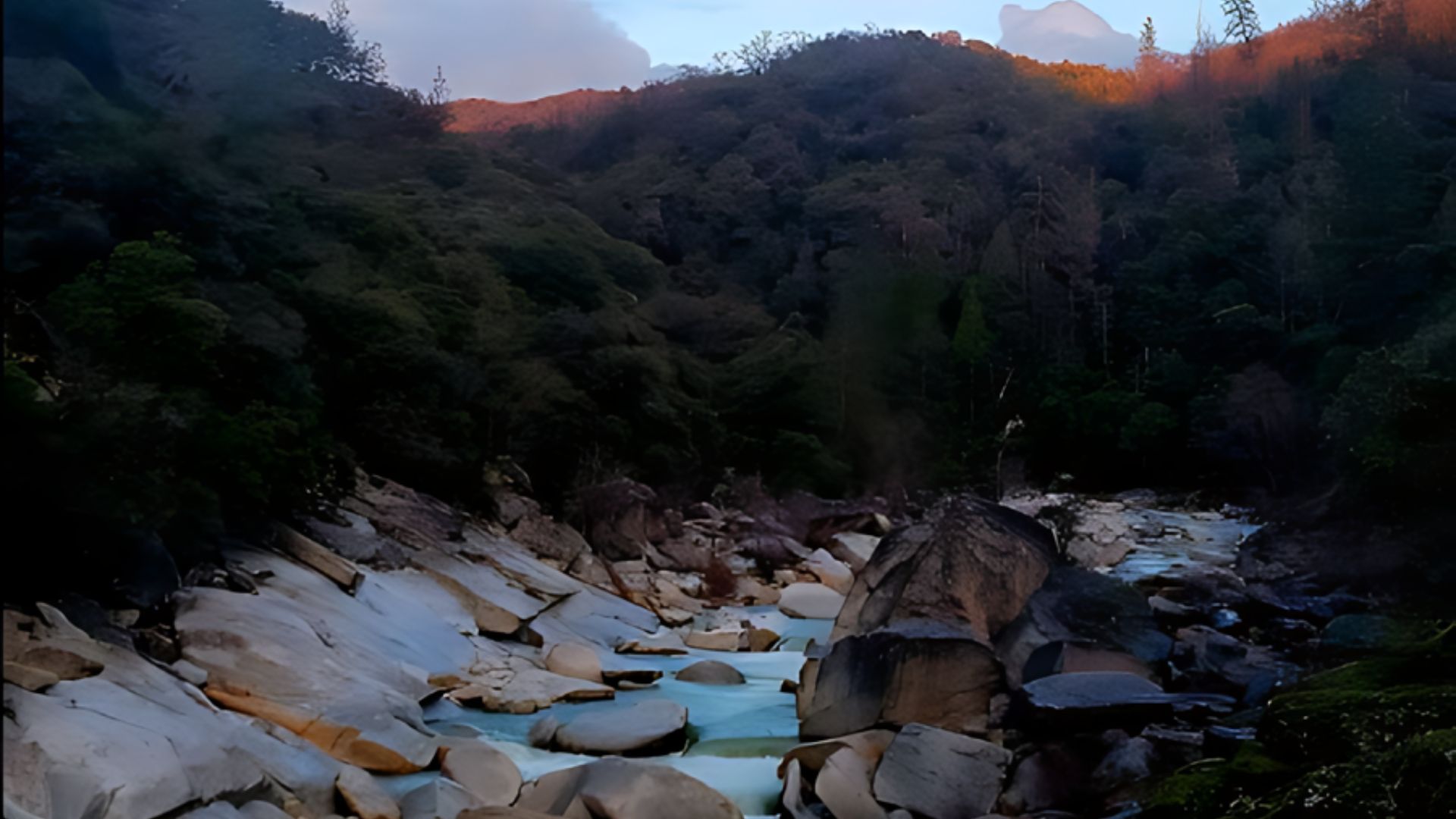 A scenic view of a rocky riverbed with a flowing river, flanked by tree-covered hills under a partly cloudy sky with a visible moon. The hills on the right show patches of warm sunlight, while the hills on the left are in shadow. The river flows over and around large boulders and smooth rock formations.