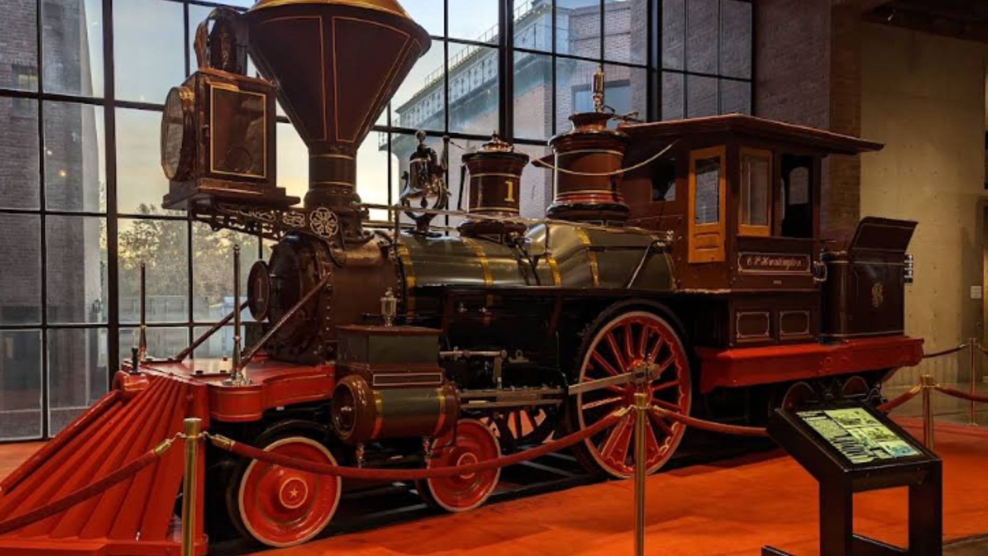 A vintage steam locomotive, the C. P. Huntington, is displayed on a red carpet in a museum setting with large windows in the background. The locomotive is dark brown and red with gold accents, featuring prominent wheels, a large smokestack, and a detailed cab.