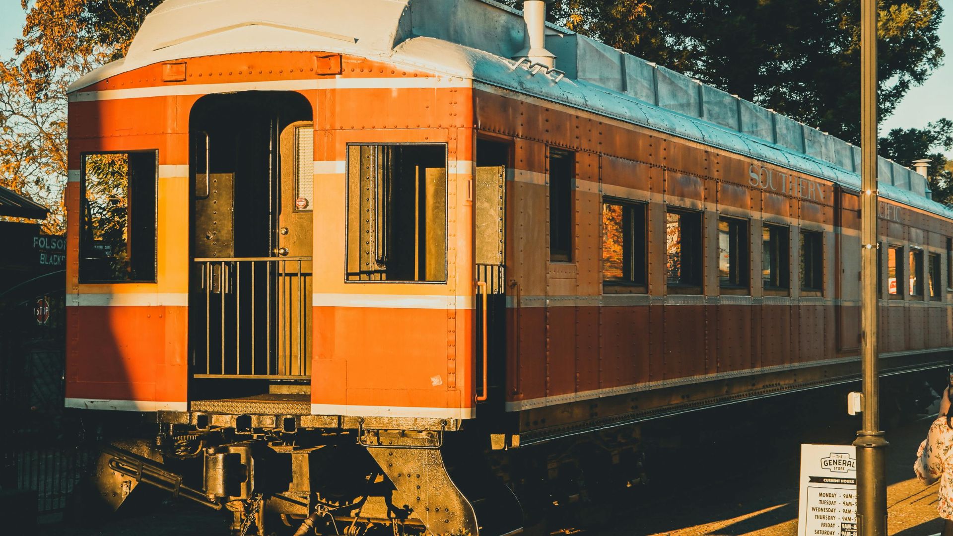A vintage orange and white train car, possibly a passenger car, sits on tracks under a warm, golden light with trees in the background. The car has multiple windows and an open doorway with a railing at one end, suggesting a classic or historical railway setting.