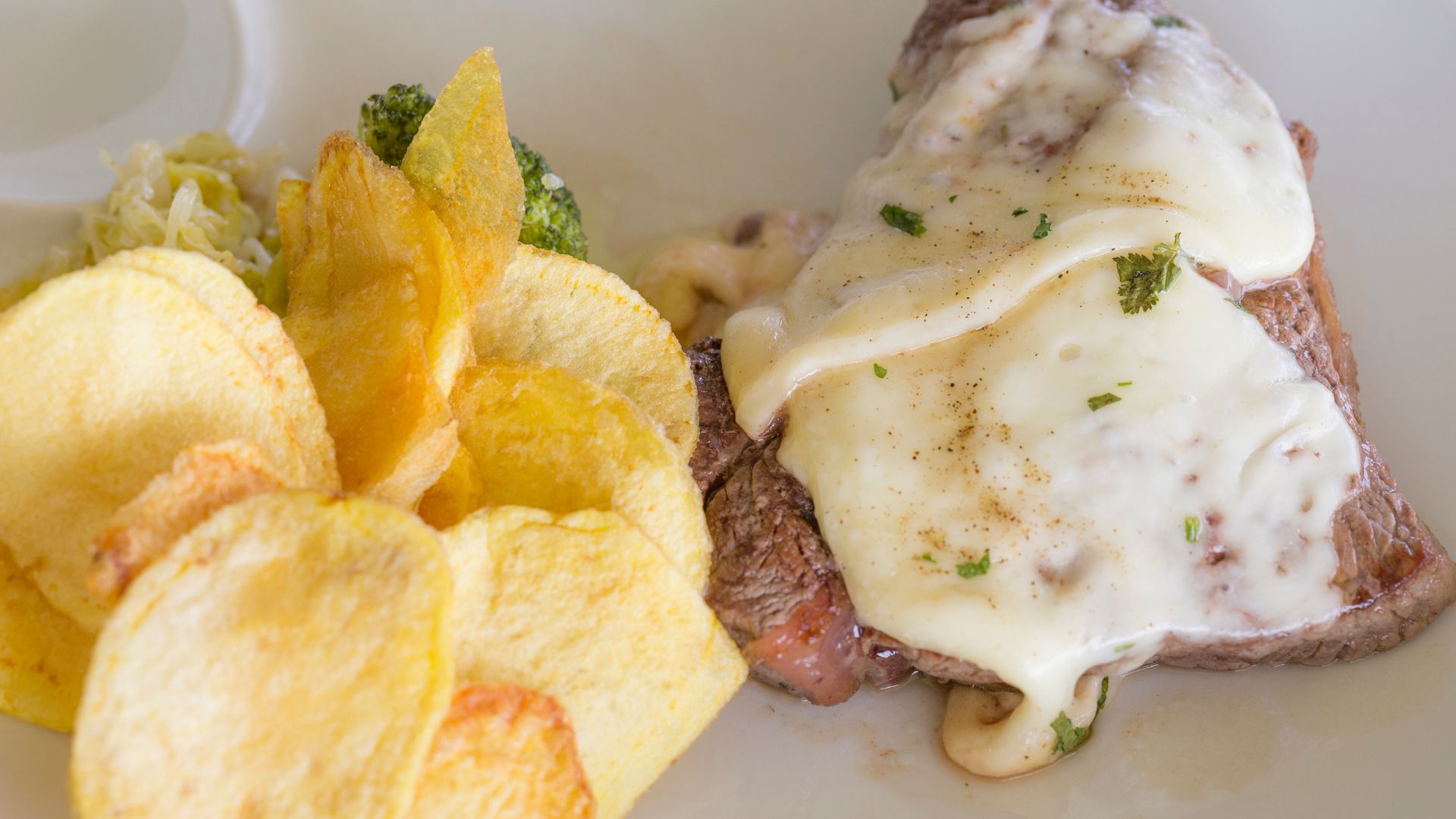 A close-up of steak topped with melted cheese, served with potato chips and a side of green vegetables.