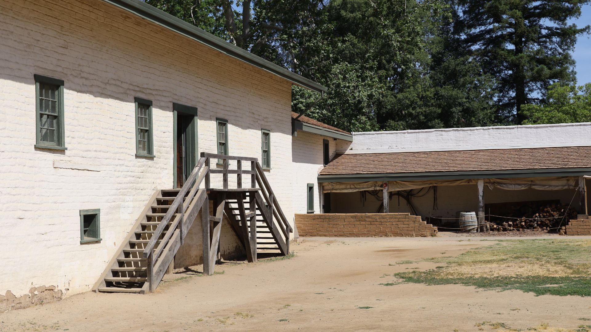 A two-story, off-white adobe building with a brown roof and green-framed windows, featuring a prominent wooden staircase leading to the second floor. To the right, a single-story structure with a similar brown roof and an open, sheltered area is visible, with wooden barrels and stacked logs underneath. The foreground is a dirt courtyard, and trees are visible in the background under a clear sky.