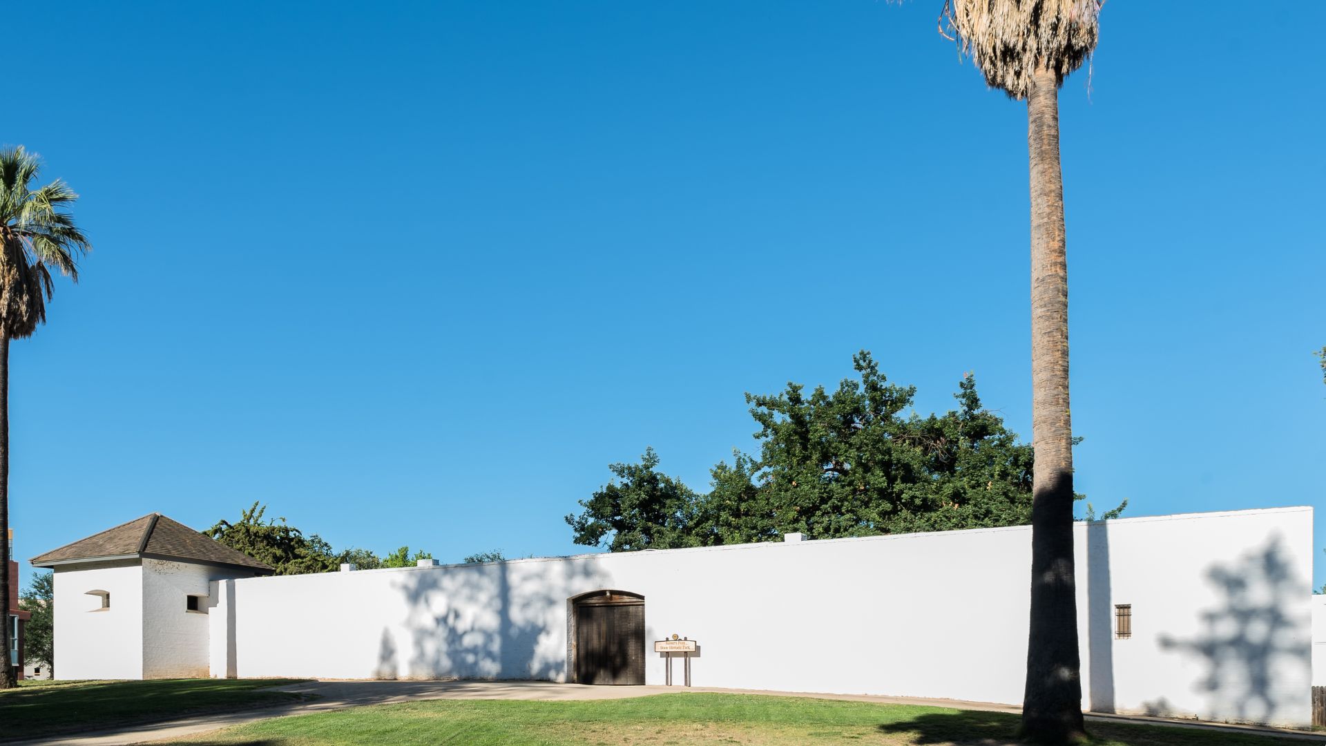 A wide shot of Sutter's Fort State Historic Park under a clear blue sky. The fort features tall, white adobe walls with a prominent wooden gate in the center, flanked by a small, white building with a brown roof on the left and a tall palm tree on the right. Green grass covers the foreground, with shadows cast by the fort and trees.