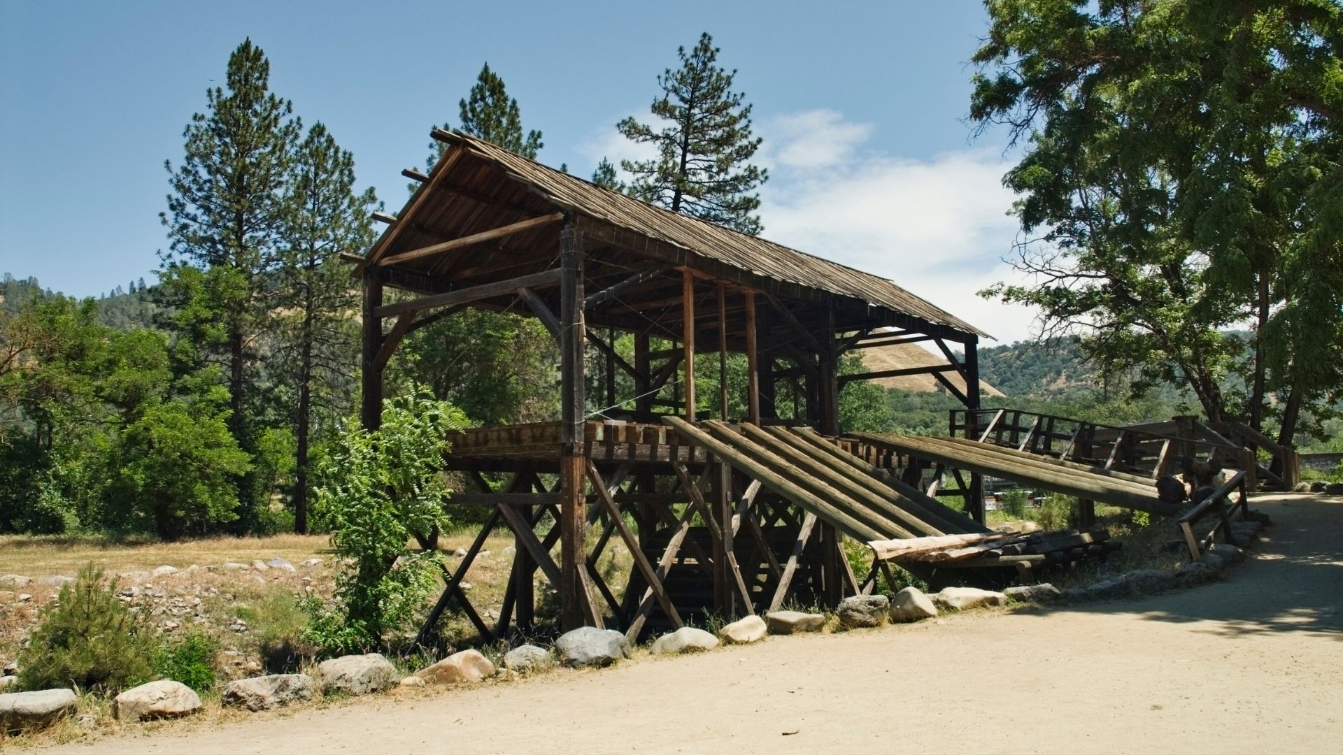 A rustic, weathered wooden covered structure, resembling a mill or flume, stands on stilts over what appears to be a dry creek bed or path. The structure has a pitched roof and is constructed with heavy timber beams and planks, showing signs of age. Lush green trees and vegetation surround the structure, and a clear blue sky with scattered clouds is visible overhead. A dirt path curves in the foreground, leading towards the structure.