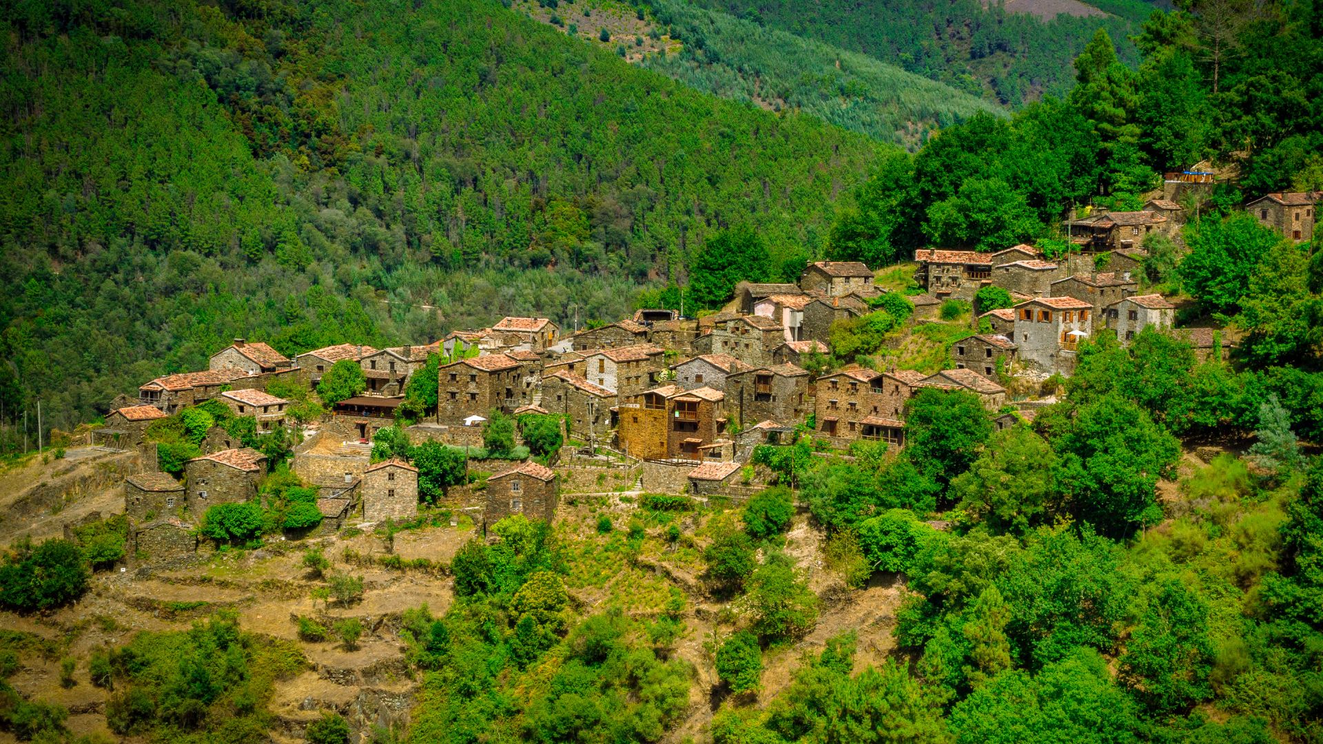A panoramic view of Linhares da Beira, a historic Portuguese village with terracotta-roofed houses nestled on a hillside, surrounded by lush green mountains under a clear blue sky.