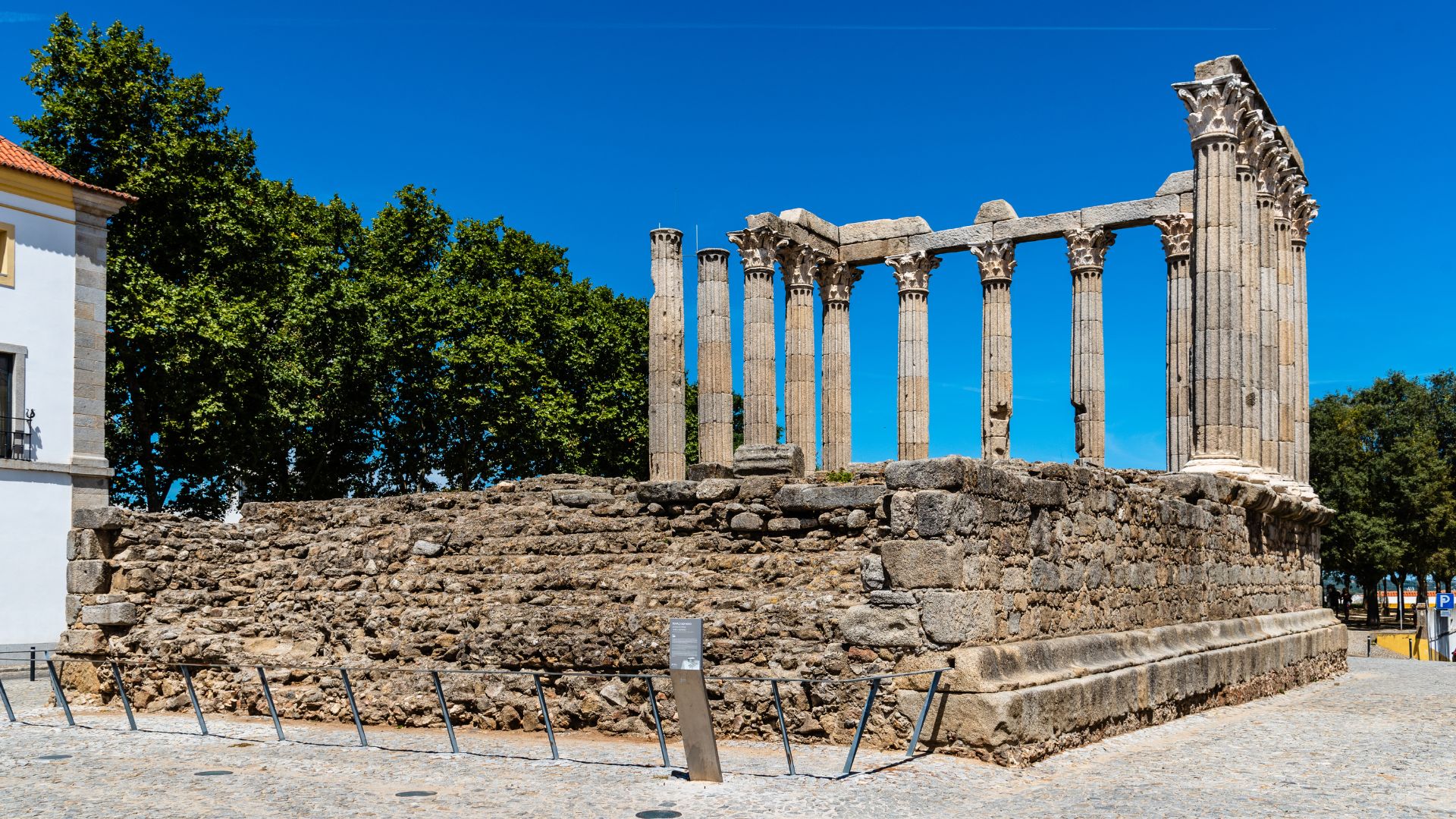 A well-preserved ancient Roman temple with tall, slender columns standing on a raised stone platform under a clear blue sky, surrounded by green trees and a modern building on the left.