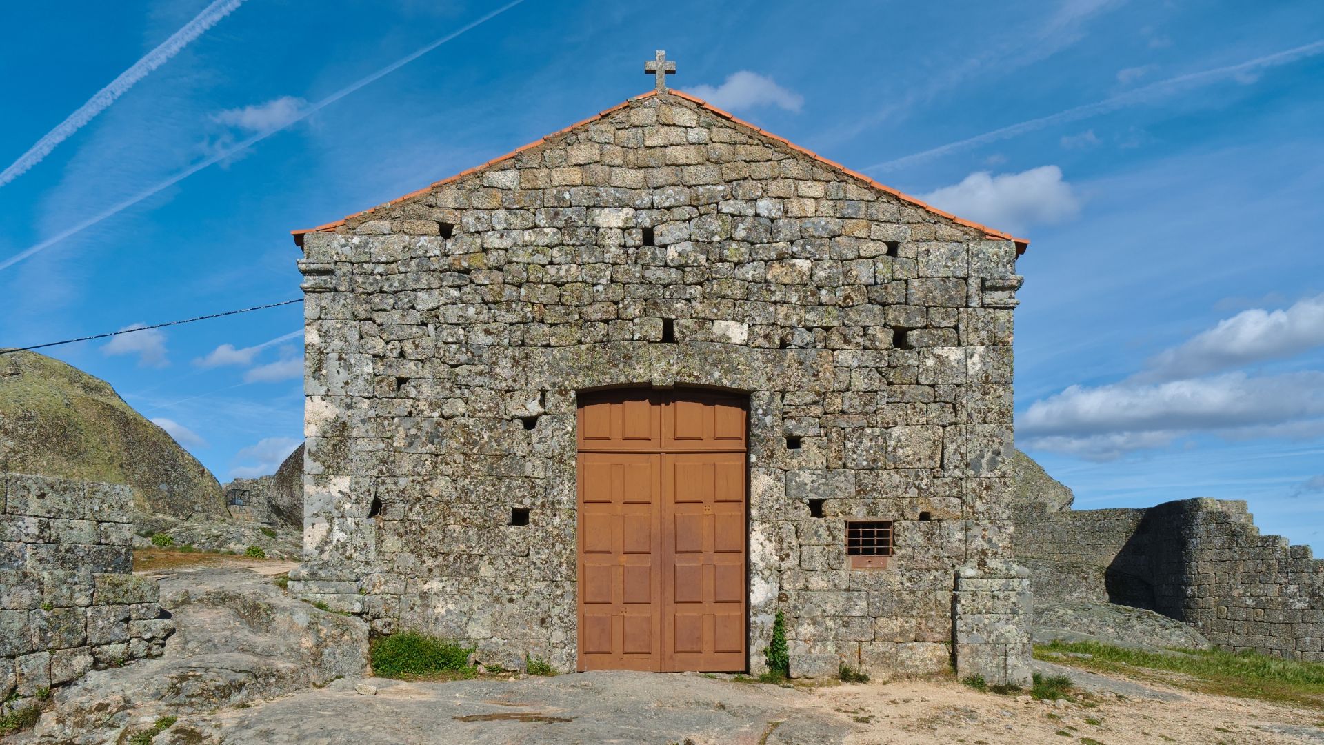 The image depicts the Chapel of Santa Maria do Castelo, a small stone chapel located within the walls of the Castle of Monsanto in Portugal. The chapel is constructed of rough-hewn stone blocks, features a brown wooden door, and has a cross atop its roof, set against a bright blue sky with wispy clouds. 