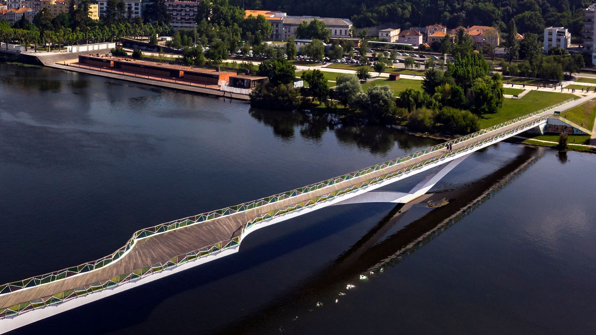 An aerial view of the modern, colorful Ponte Pedro e Inês pedestrian bridge spanning a river, with green parkland and city buildings visible on the far bank.