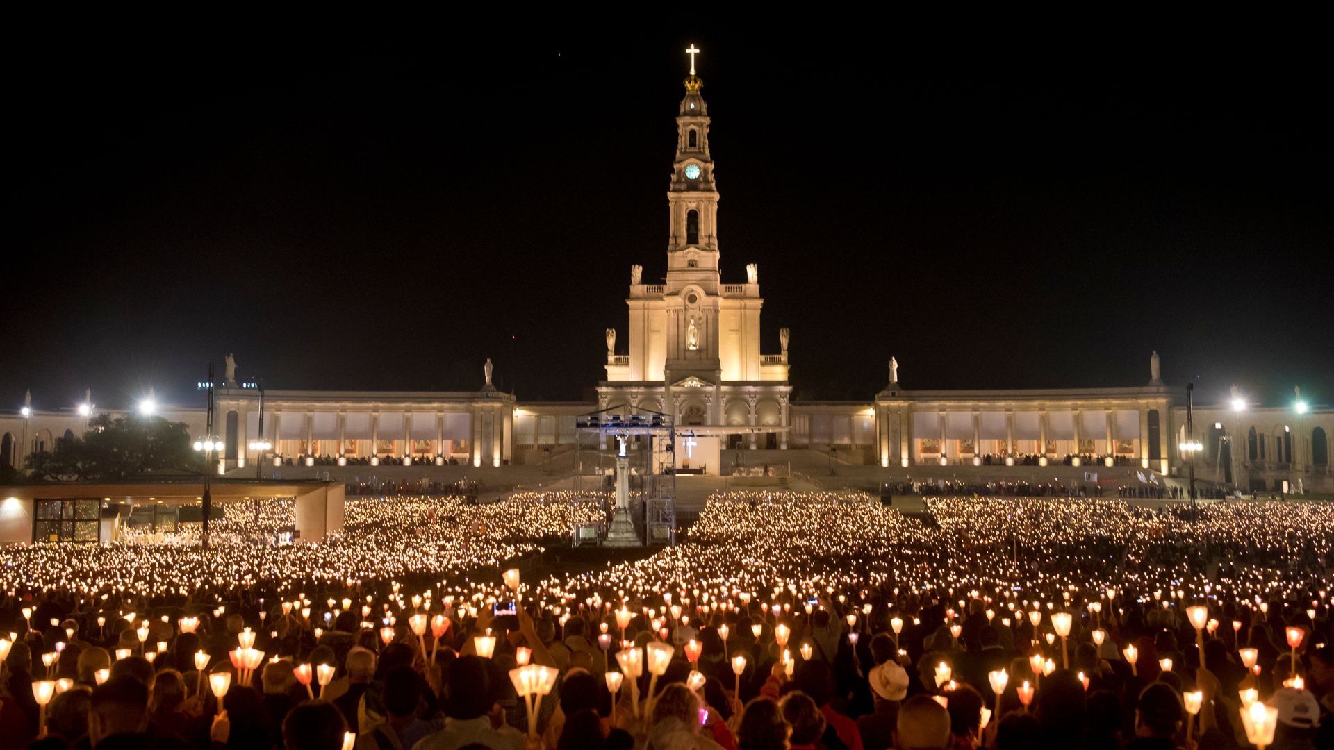 A wide shot at night of the Sanctuary of Fátima in Portugal, illuminated and surrounded by a vast crowd of pilgrims holding lit candles during a candlelight procession. 