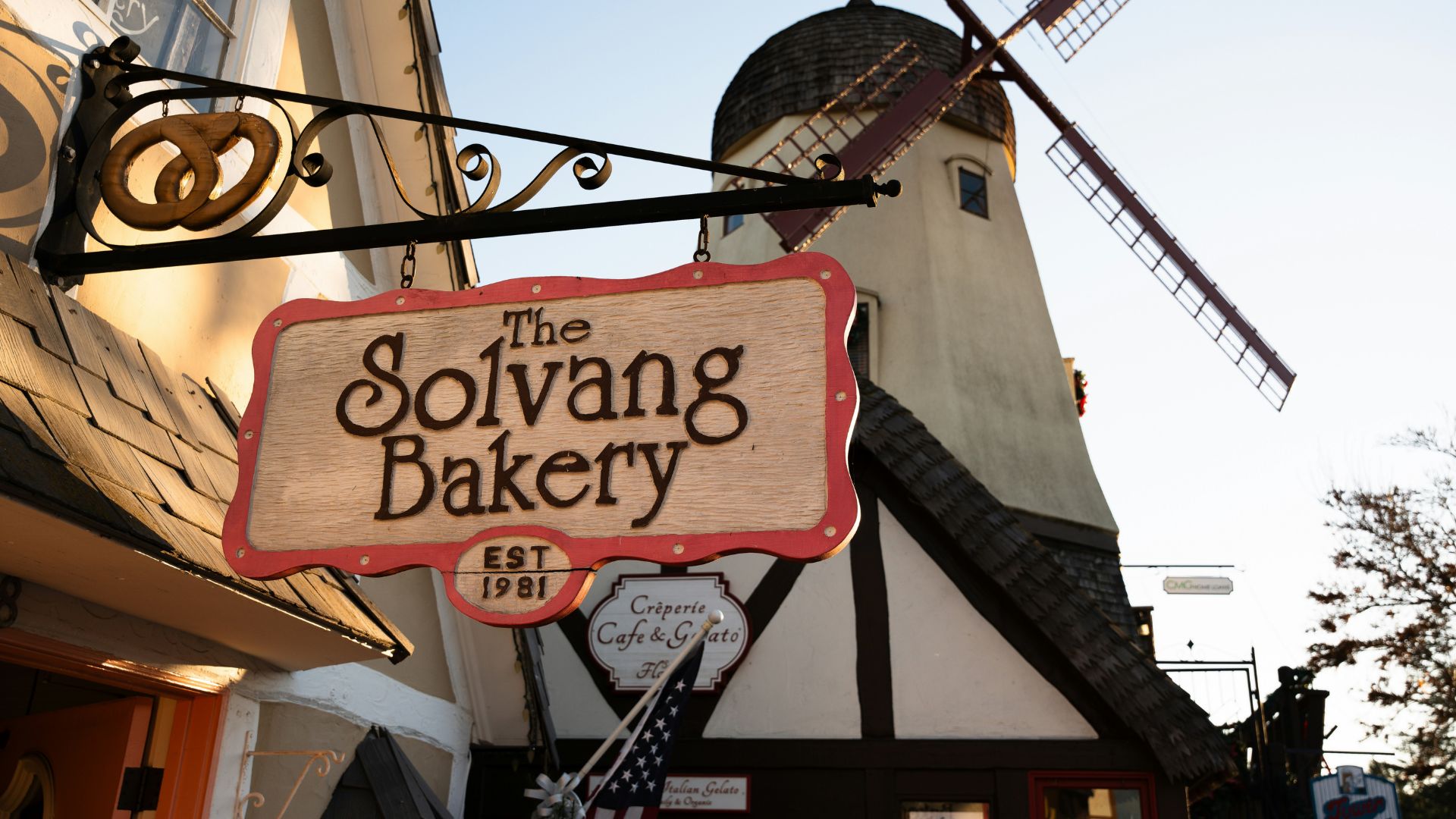  A close-up view of the sign for "The Solvang Bakery" with a traditional Danish-style windmill visible in the background against a clear sky. The sign also indicates "EST 1981" and mentions "Creperie Cafe & Gato.