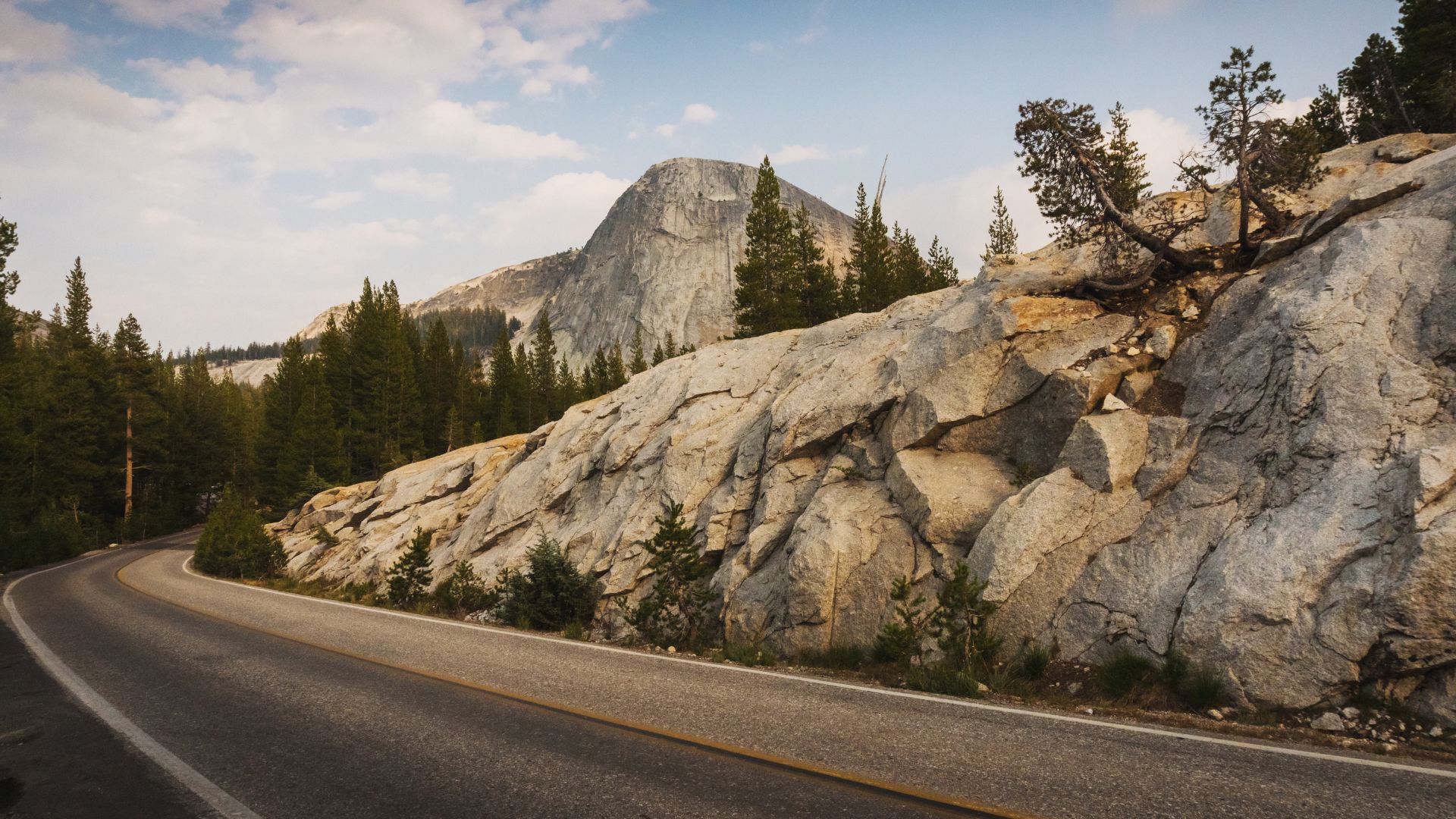 A winding road, Tioga Road (California State Route 120), curves through a mountainous landscape within Yosemite National Park, featuring large granite rock formations, scattered pine trees, and a prominent peak in the background under a partly cloudy sky. 