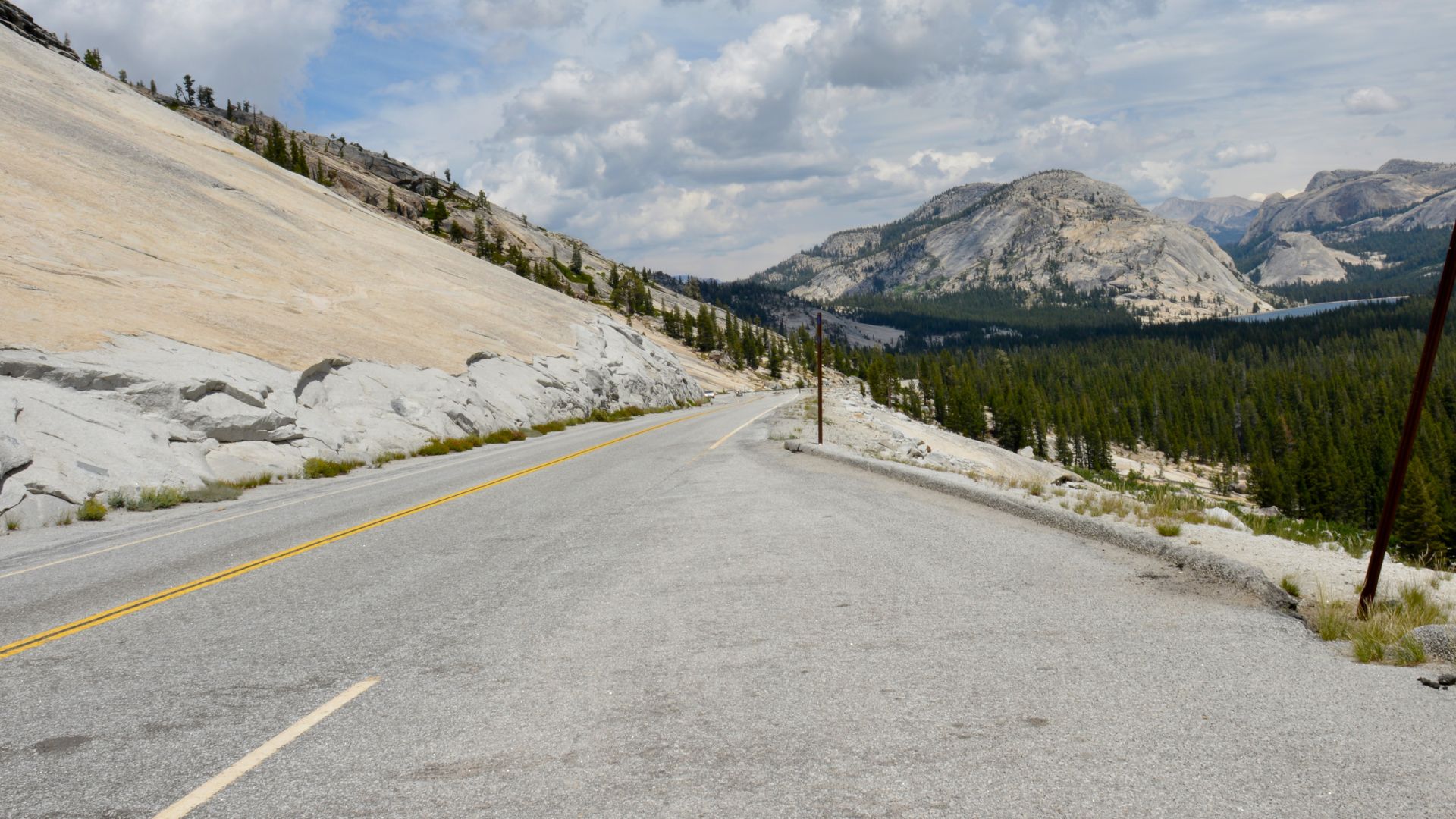 A scenic view of Tioga Road winding through Tioga Pass in Yosemite National Park, with granite domes and evergreen trees on either side and mountains in the distance under a cloudy sky.