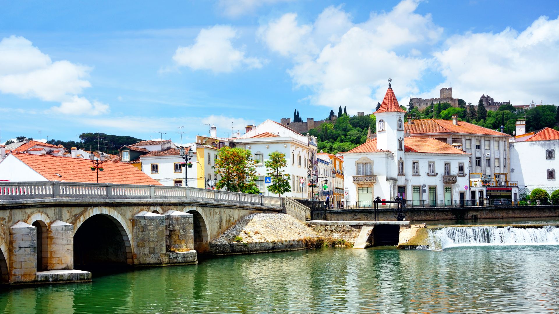 A vibrant image of the historic city of Tomar, Portugal, showing an arched stone bridge over a river, with colorful buildings lining the riverbanks, and a prominent castle and convent complex perched atop a hill in the background under a blue sky with white clouds.