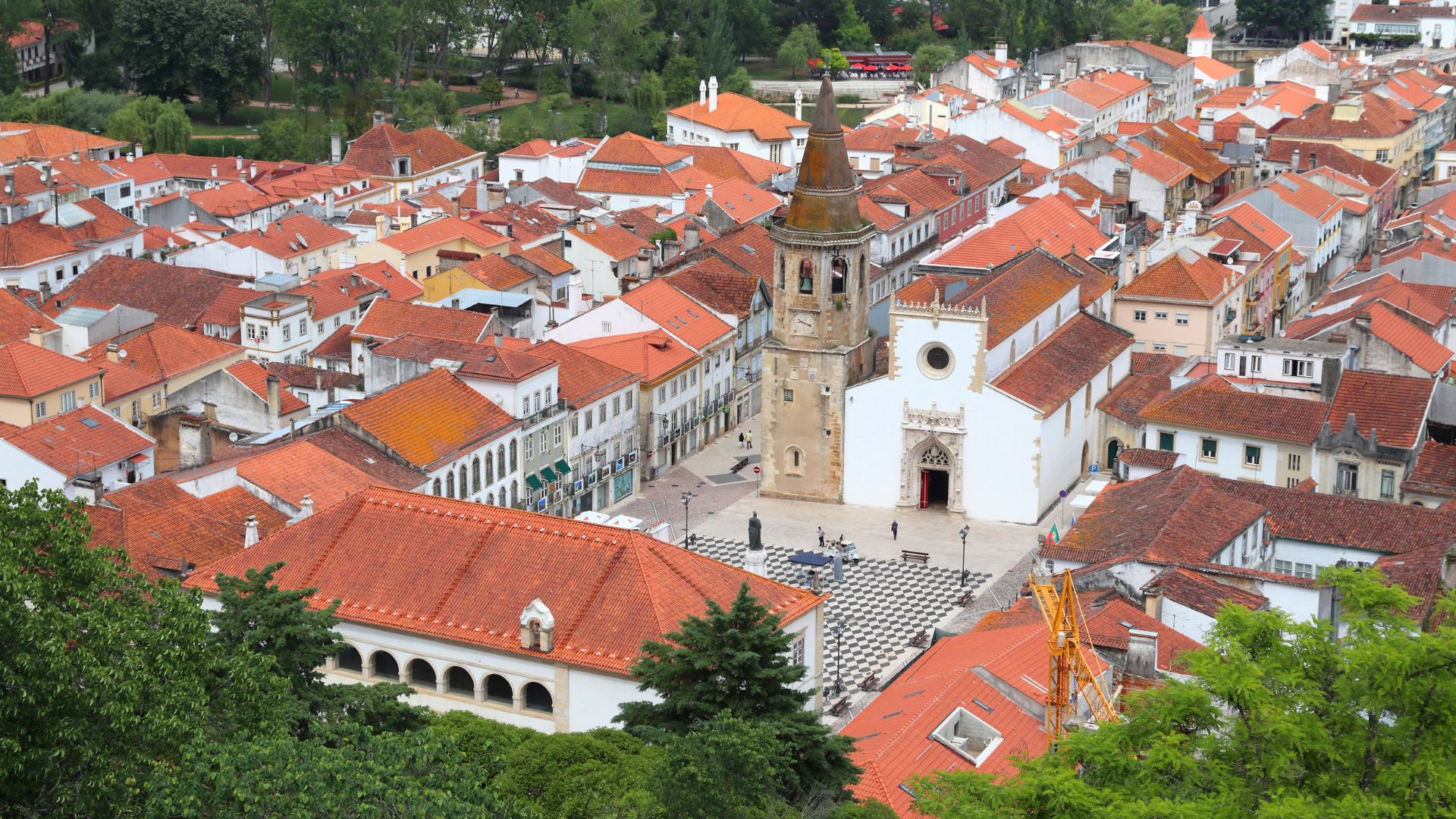 An aerial view of the historic city of Tomar in central Portugal, showcasing its dense urban landscape with numerous orange-tiled roofs, a prominent white church (Igreja de São João Baptista) with a bell tower at its center, and surrounding green trees.