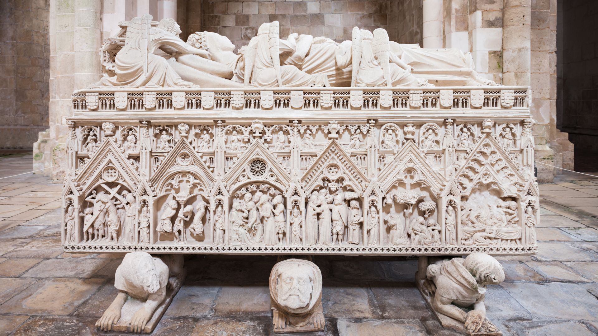A detailed image of the elaborately carved white marble tomb of Inês de Castro, featuring sculpted figures on the lid and intricate relief scenes from the New Testament along its sides, resting on six hybrid figures with human faces and beast bodies.