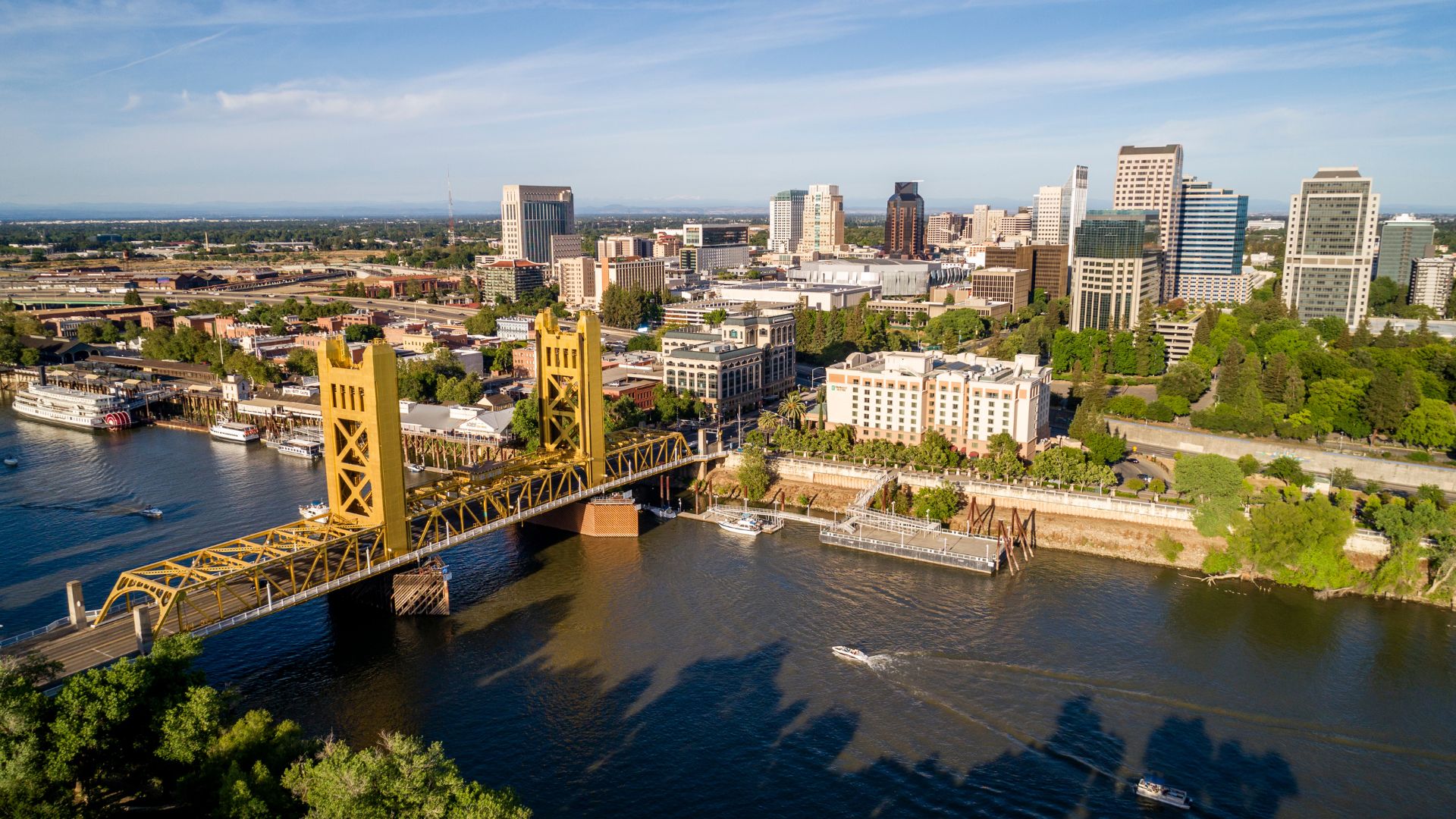 An aerial view of the Tower Bridge, a golden yellow vertical lift bridge, spanning the Sacramento River, with the downtown Sacramento skyline visible in the background under a clear sky. Boats are visible on the river, and lush green trees line the riverbanks.