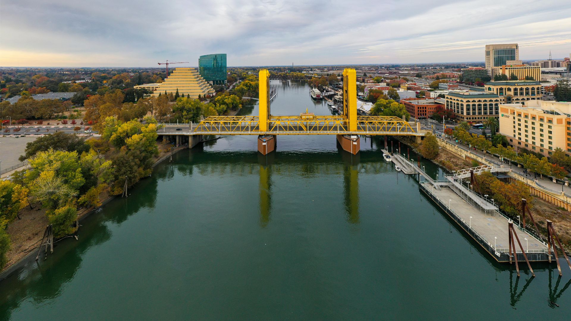 An aerial view captures the vibrant yellow Tower Bridge spanning a calm, dark green river under a cloudy sky. The bridge connects a tree-lined shore with buildings on the left to a developed riverfront with structures and a pier on the right. In the background, the cityscape of Sacramento is visible, including a distinctive pyramid-shaped building and other urban structures.