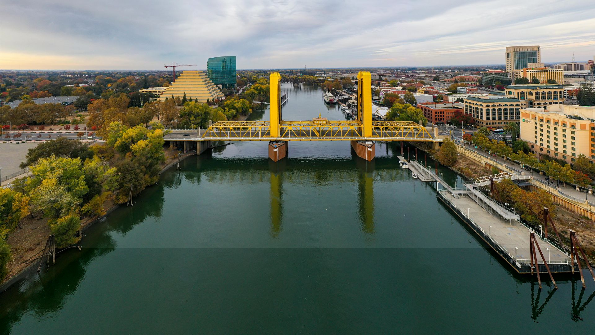 An aerial view captures the vibrant yellow Tower Bridge spanning a calm, dark green river under a cloudy sky. The bridge connects a tree-lined shore with buildings on the left to a developed riverfront with structures and a pier on the right. In the background, the cityscape of Sacramento is visible, including a distinctive pyramid-shaped building and other urban structures.