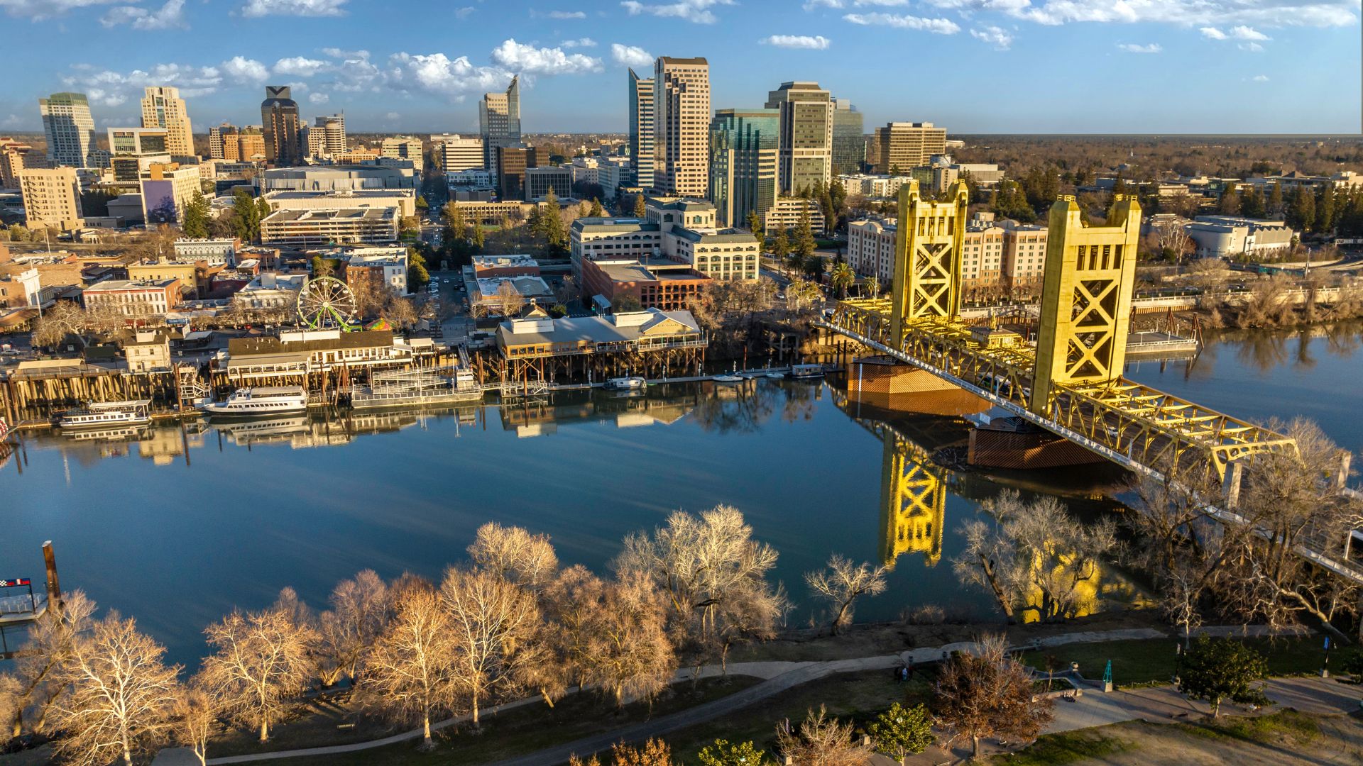 An aerial view of the Sacramento, California skyline at sunset, with the prominent yellow Tower Bridge crossing the Sacramento River in the foreground. The city's buildings are reflected in the calm river, and bare trees line the riverbanks, suggesting a cooler season.