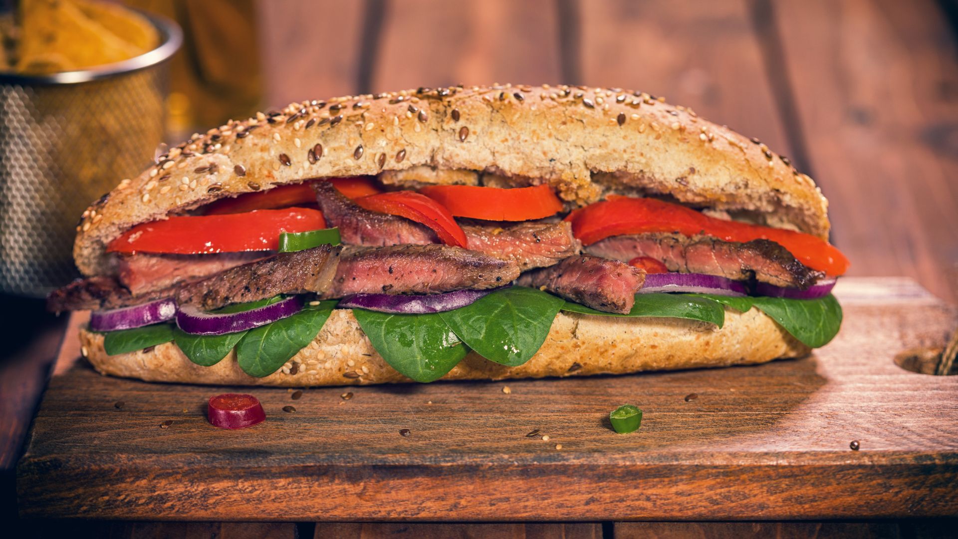 A close-up shot of a hearty steak sandwich on a seeded baguette, filled with sliced meat (likely tri-tip), red and green bell peppers, red onions, and spinach leaves, presented on a wooden cutting board.