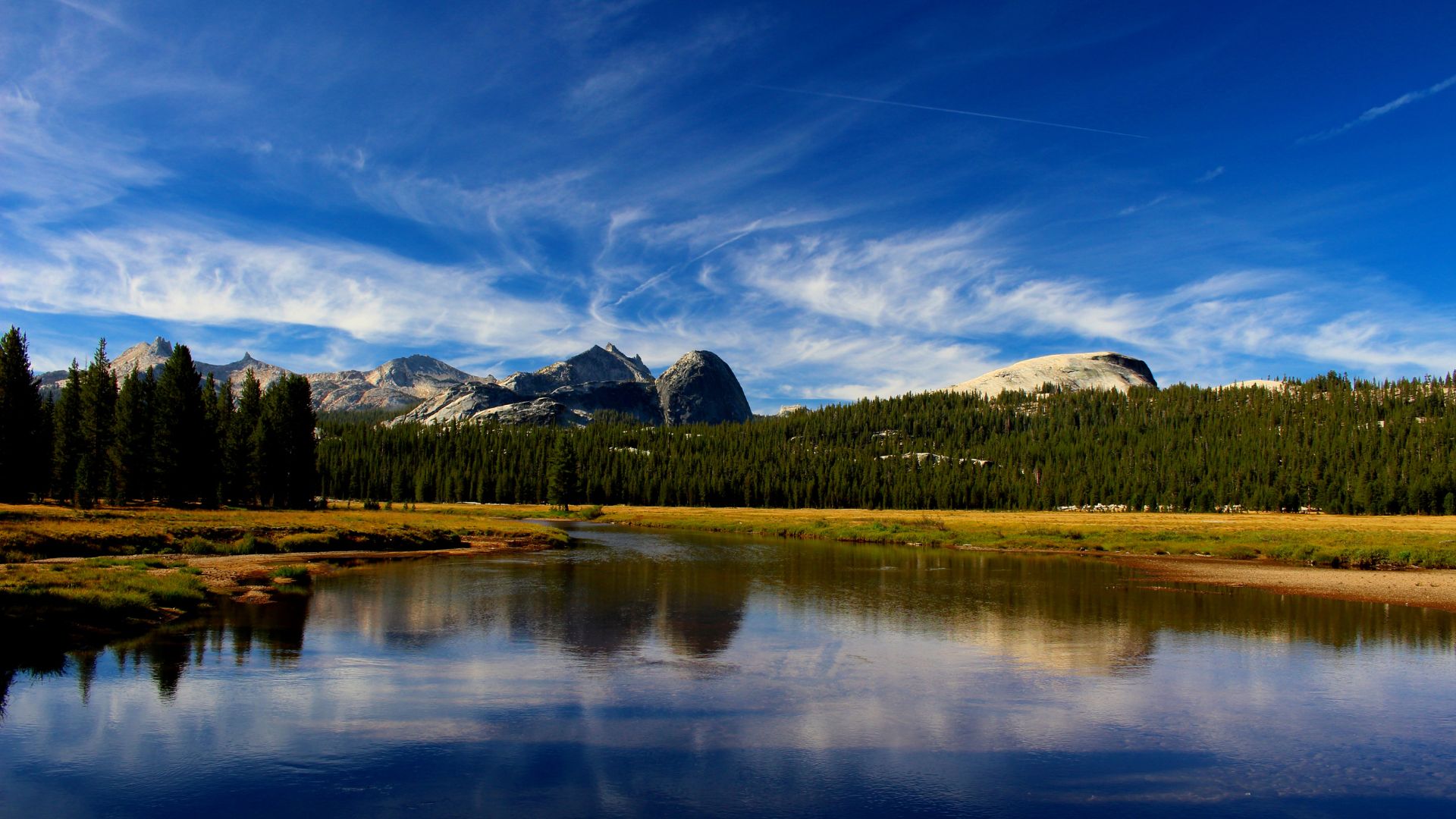 A wide shot of a calm river reflecting a clear blue sky with white clouds, flanked by lush green forests and towering granite mountains in the background, under a bright, sunny day.