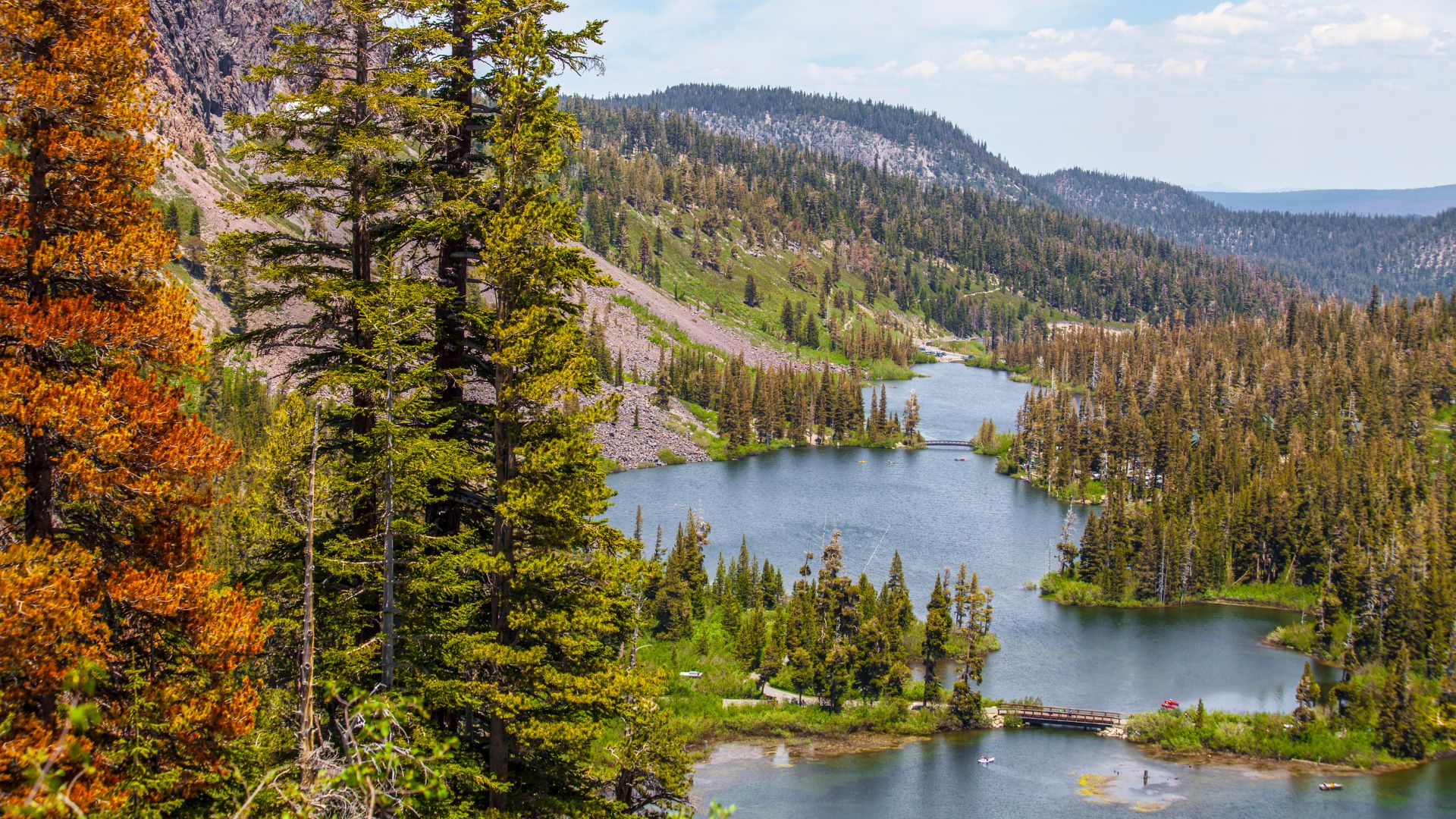 A scenic landscape view of Twin Lakes in Mammoth Lakes, California, featuring two interconnected lakes surrounded by dense evergreen and some autumn-colored trees, with a mountainous backdrop under a partly cloudy sky. A small bridge spans the narrow section between the two lakes, and a few small boats are visible on the water. 