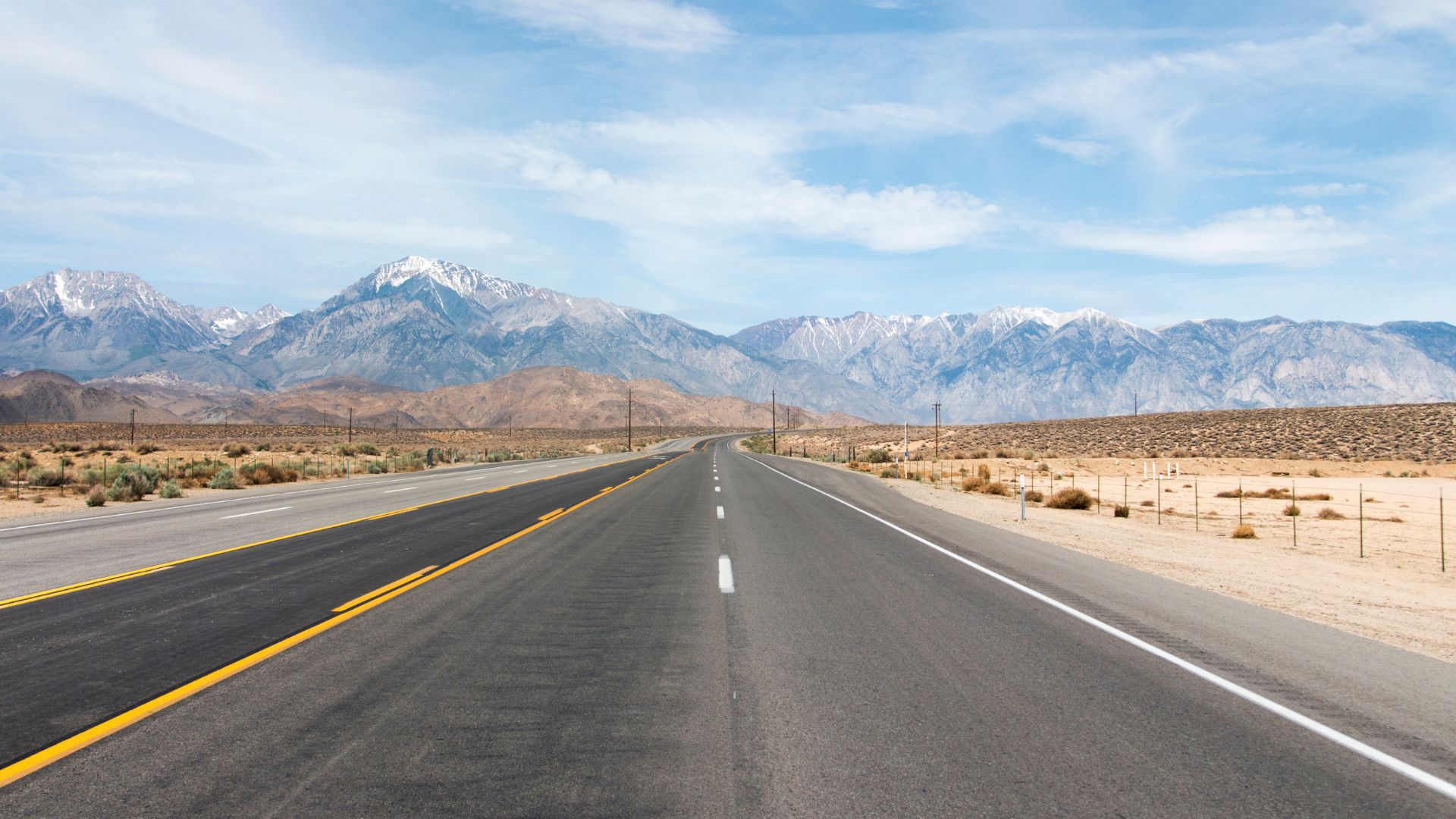 A long, empty highway (U.S. Route 395) stretches towards a range of majestic, snow-capped mountains under a clear sky in the Eastern Sierra region of California.