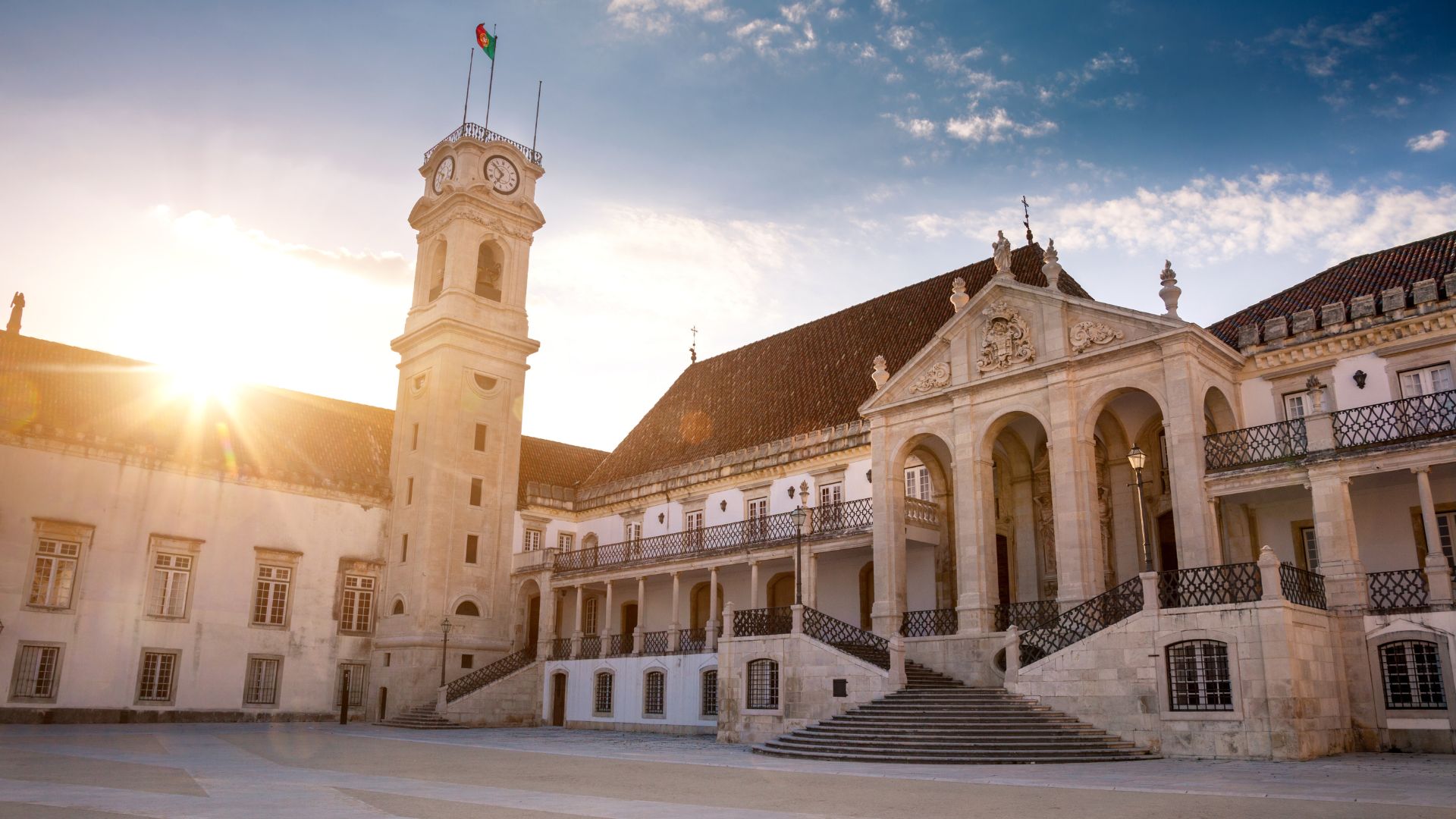 A grand, historic building with a prominent clock tower and ornate architecture, bathed in sunlight, representing the University of Coimbra in Portugal.