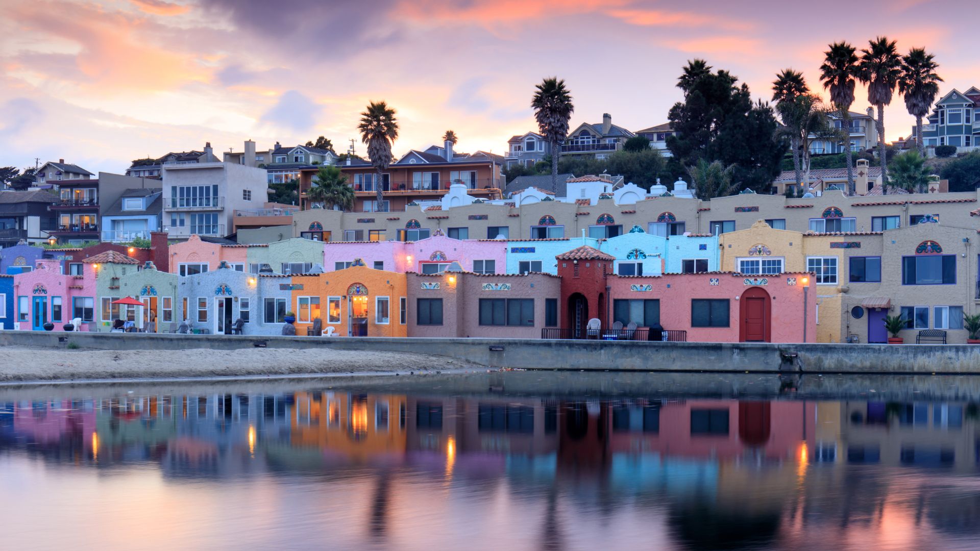 An eye-level shot captures the colorful Venetian Court condominiums and the Capitola Venetian Hotel reflecting in the calm waters of the Capitola Lagoon at sunset. The pastel-colored buildings, ranging from pinks and oranges to blues and greens, line the waterfront, with palm trees and additional houses visible on the hillside above under a dramatic sky with hues of orange and purple. 