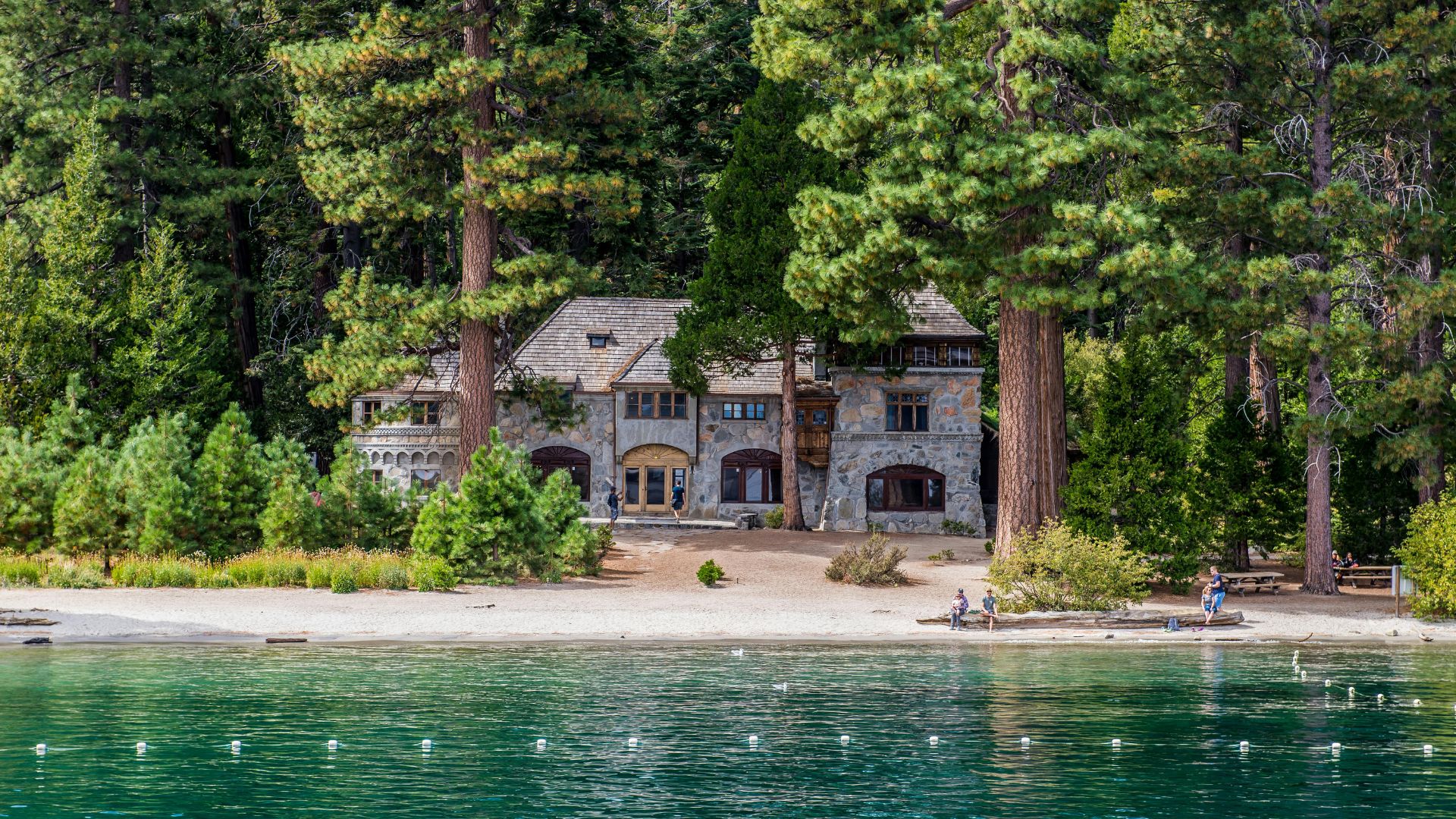 A large stone mansion with a distinctive Scandinavian architectural style, known as Vikingsholm, stands amidst tall pine trees on the sandy shore of Emerald Bay, Lake Tahoe, with the clear turquoise waters of the bay in the foreground and a few people visible on the beach.
