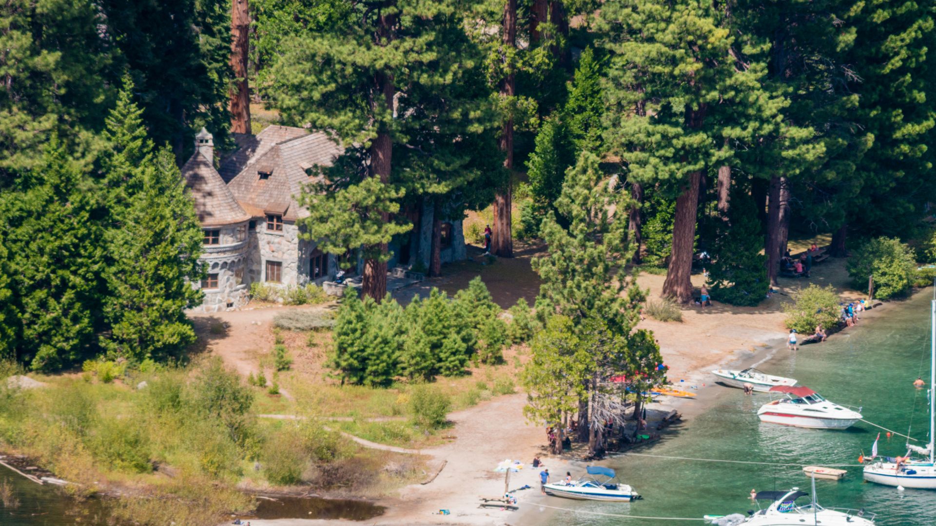 A stone and timber mansion with a sod roof, Vikingsholm, nestled among tall pine trees on the shore of Emerald Bay, Lake Tahoe, with boats visible in the clear water and a sandy beach in the foreground.