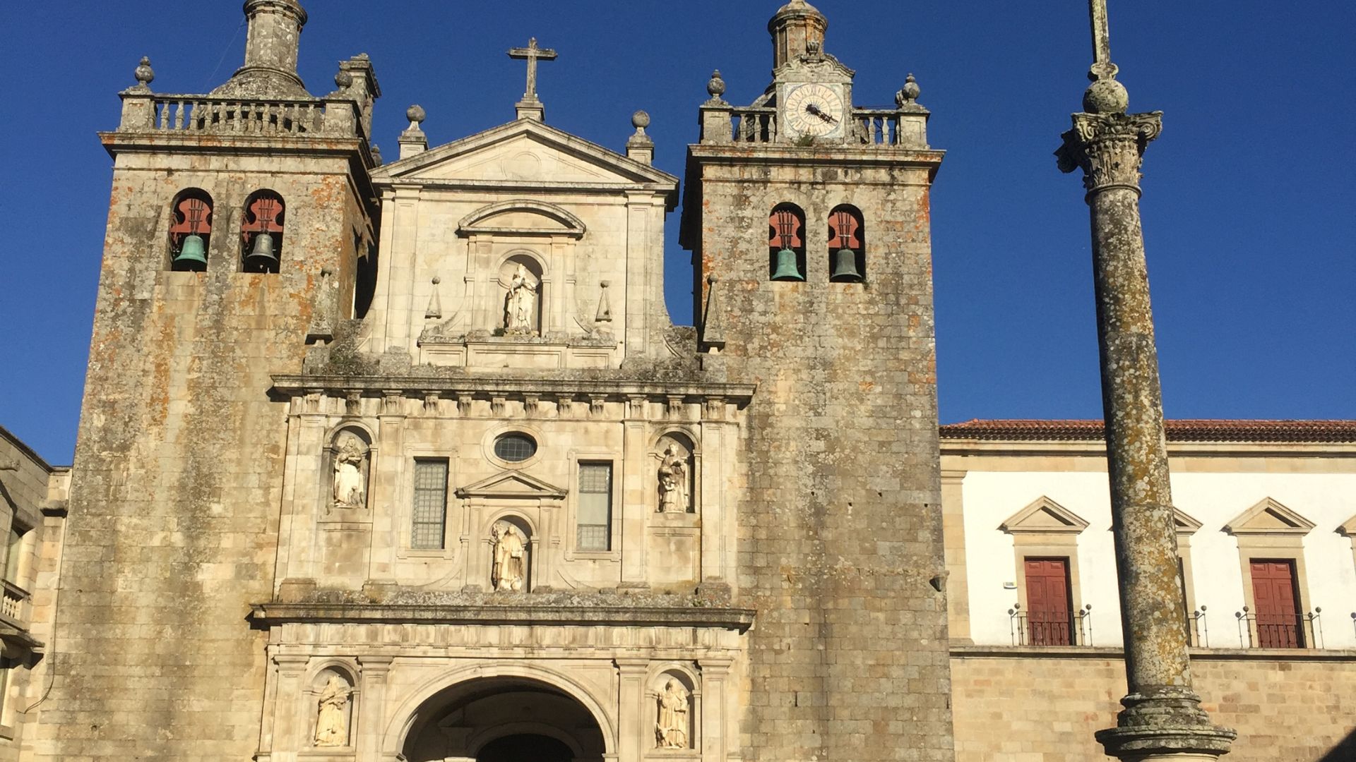 A frontal view of the Viseu Cathedral in central Portugal, showcasing its stone facade with prominent bell towers, a central cross, and various architectural details under a clear blue sky.