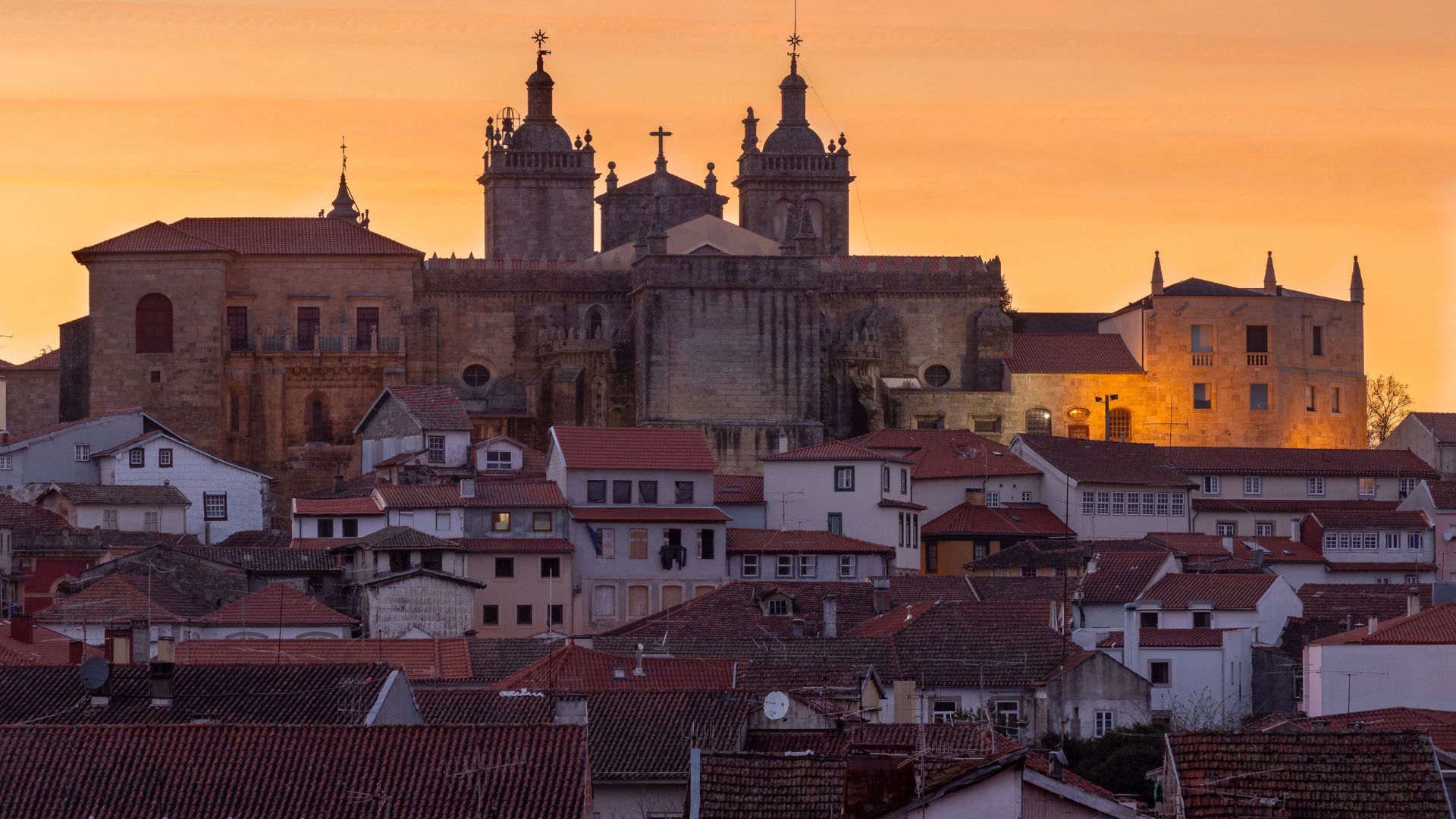 A wide shot showcasing the historic city of Viseu, Portugal, with the grand Viseu Cathedral and its distinctive twin bell towers dominating the skyline amidst a dense cluster of traditional buildings with terracotta roofs, under sunset.