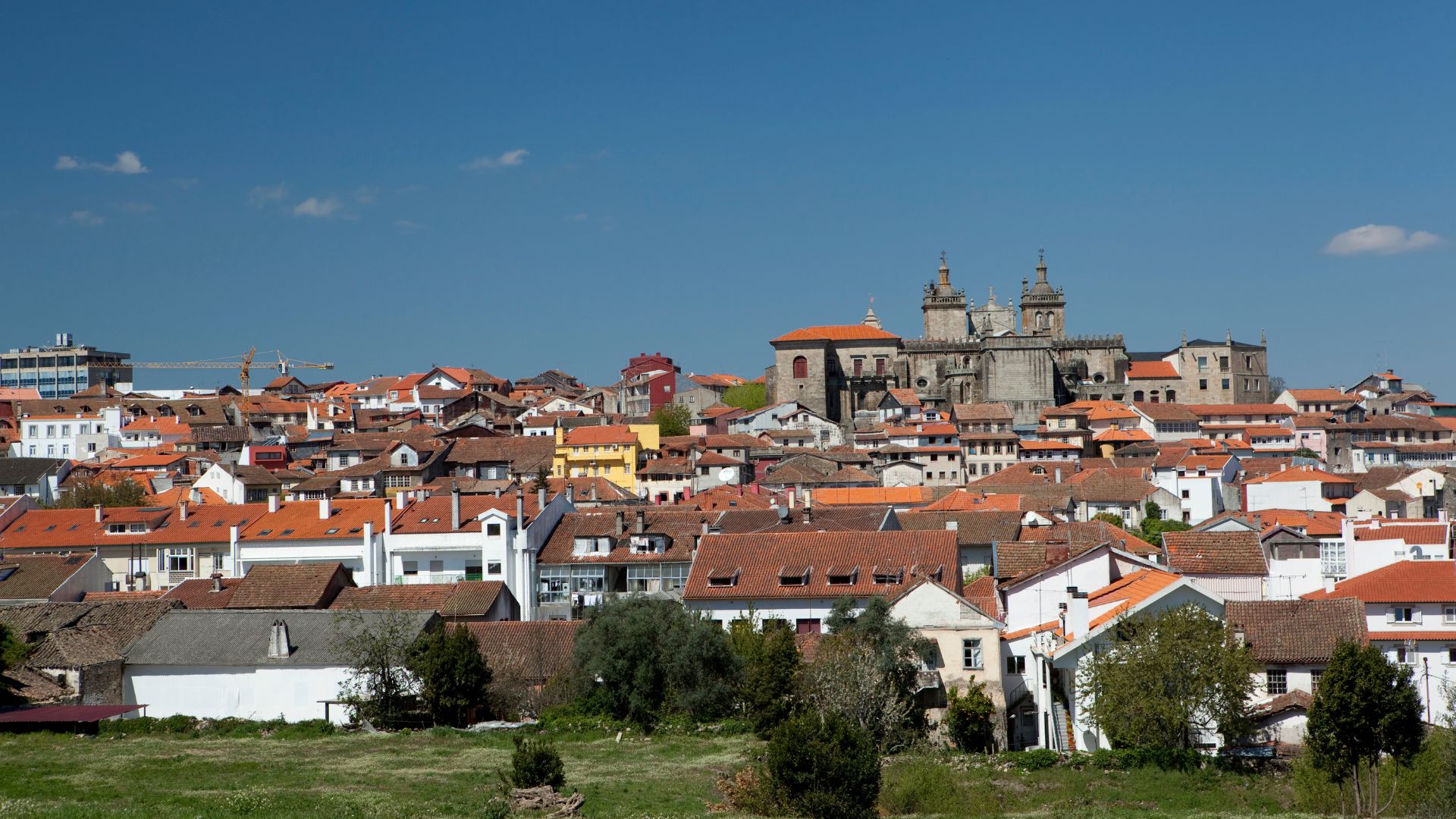 A wide shot showcasing the historic city of Viseu, Portugal, with the grand Viseu Cathedral and its distinctive twin bell towers dominating the skyline amidst a dense cluster of traditional buildings with terracotta roofs, all under a clear blue sky. 
