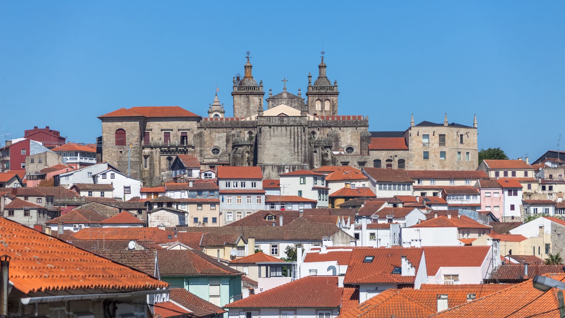 A wide shot showcasing the historic city of Viseu, Portugal, with the grand Viseu Cathedral and its distinctive twin bell towers dominating the skyline amidst a dense cluster of traditional buildings with terracotta roofs, all under a clear blue sky.