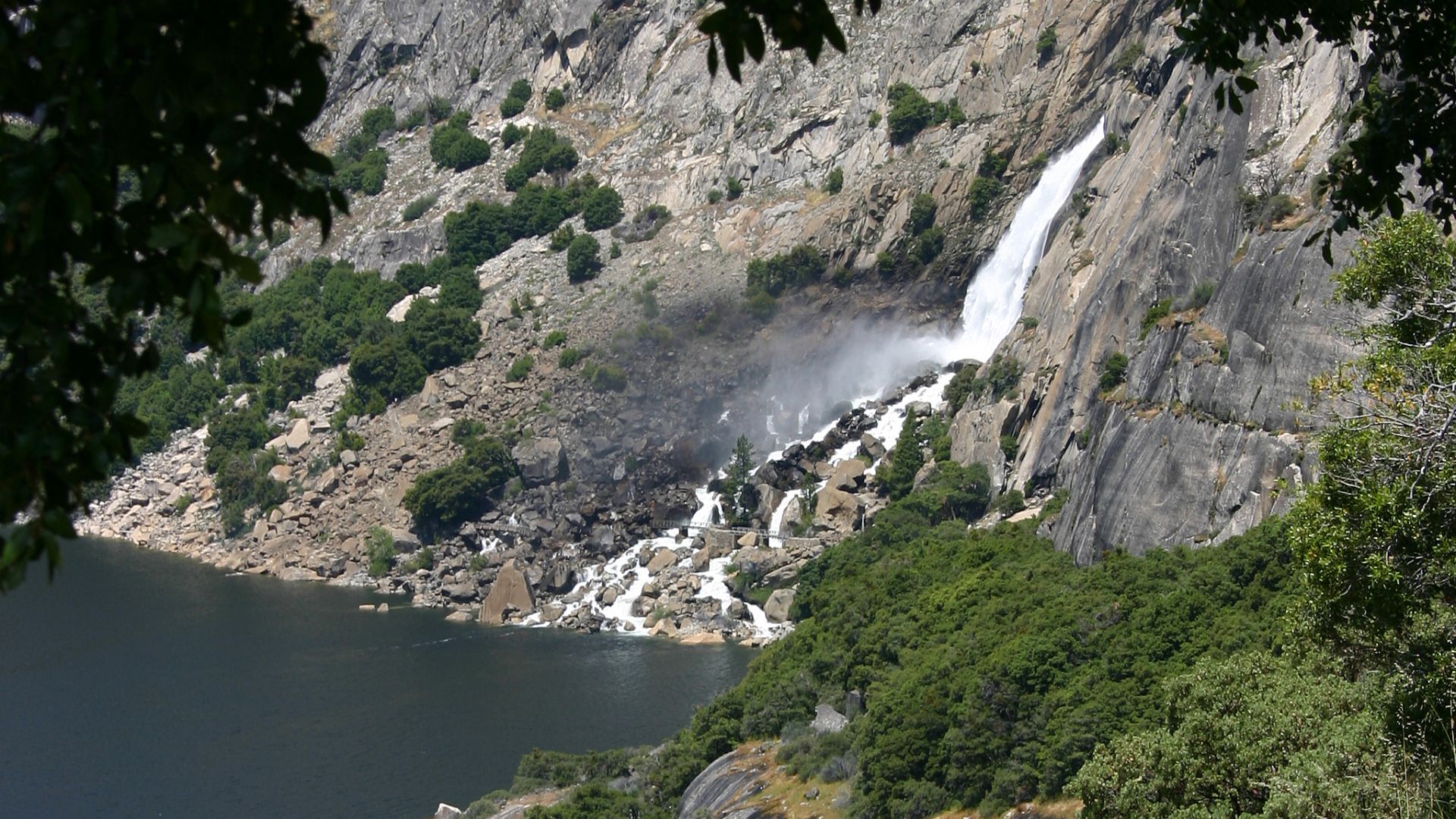 A powerful waterfall cascades down a steep, rocky cliff face into a serene lake surrounded by lush green trees and rocky terrain under a clear sky. The waterfall is prominent on the right side of the image, with the lake and surrounding landscape filling the foreground and left side.