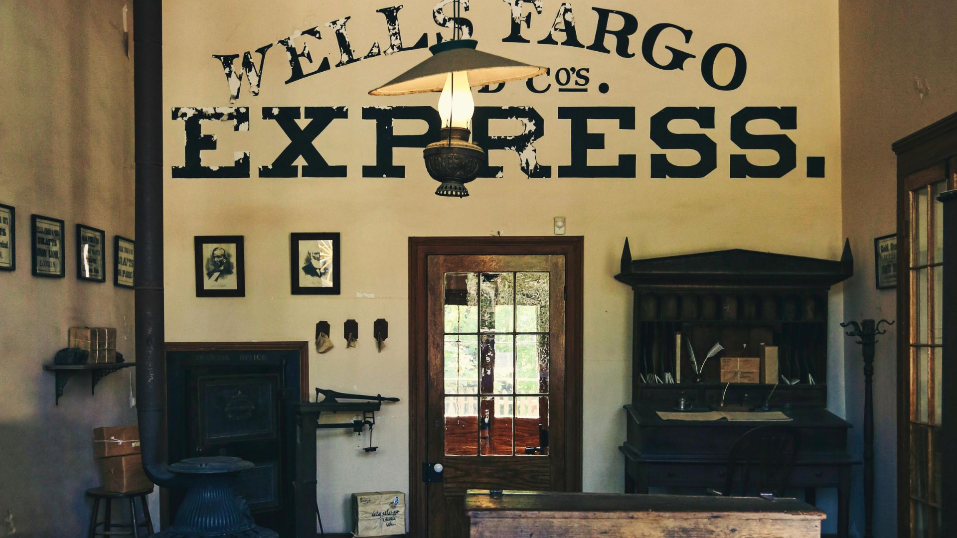 A vintage interior shot of a Wells Fargo Express office, featuring dark wooden counters and furniture, with a prominent "WELLS FARGO CO. EXPRESS." sign painted in black on a light-colored wall above a central doorway. A hanging light fixture illuminates the space.