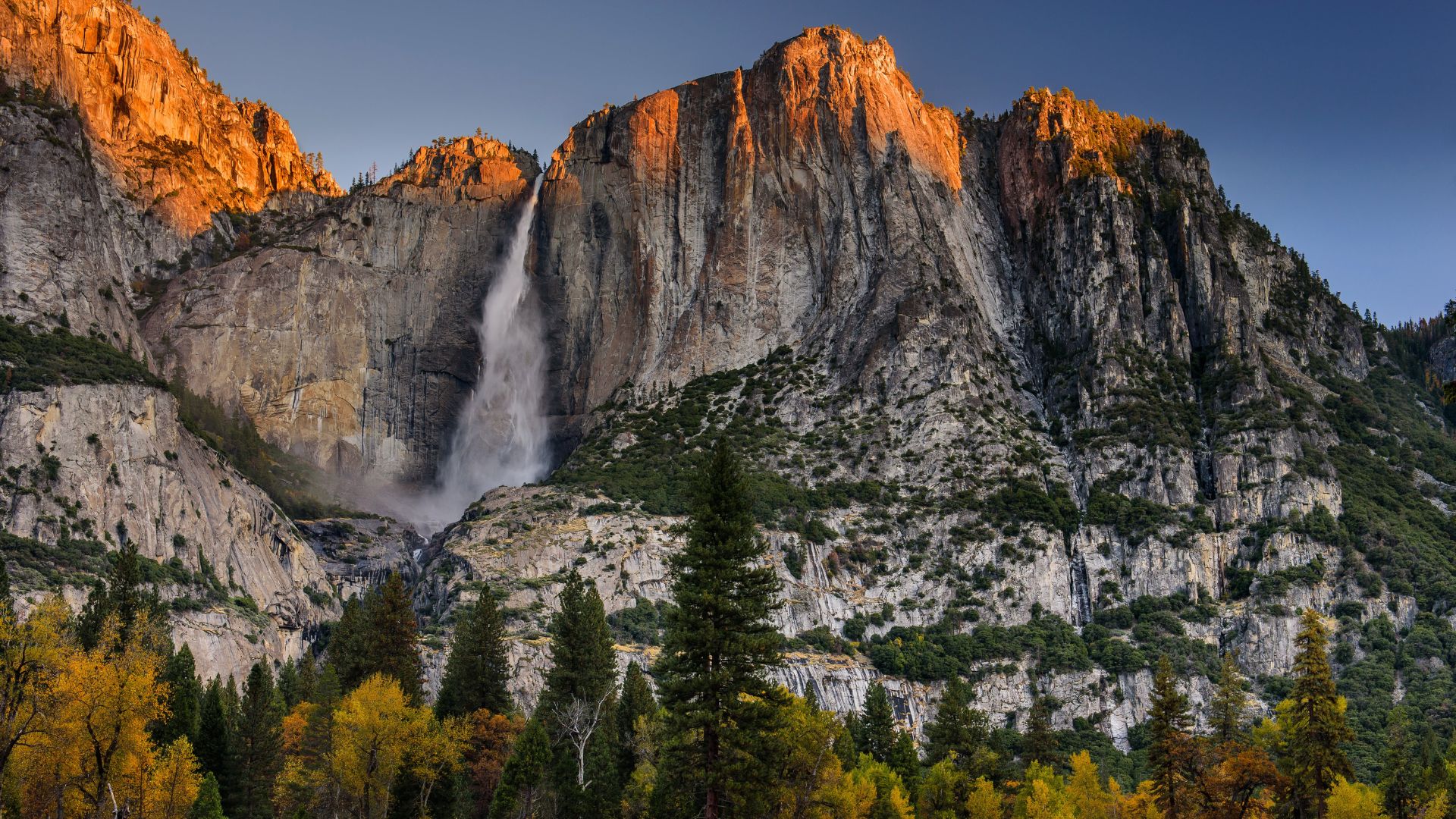 A majestic waterfall, Yosemite Falls, plunges down a massive granite cliff face, surrounded by autumn-colored trees and rugged mountains under a clear sky.