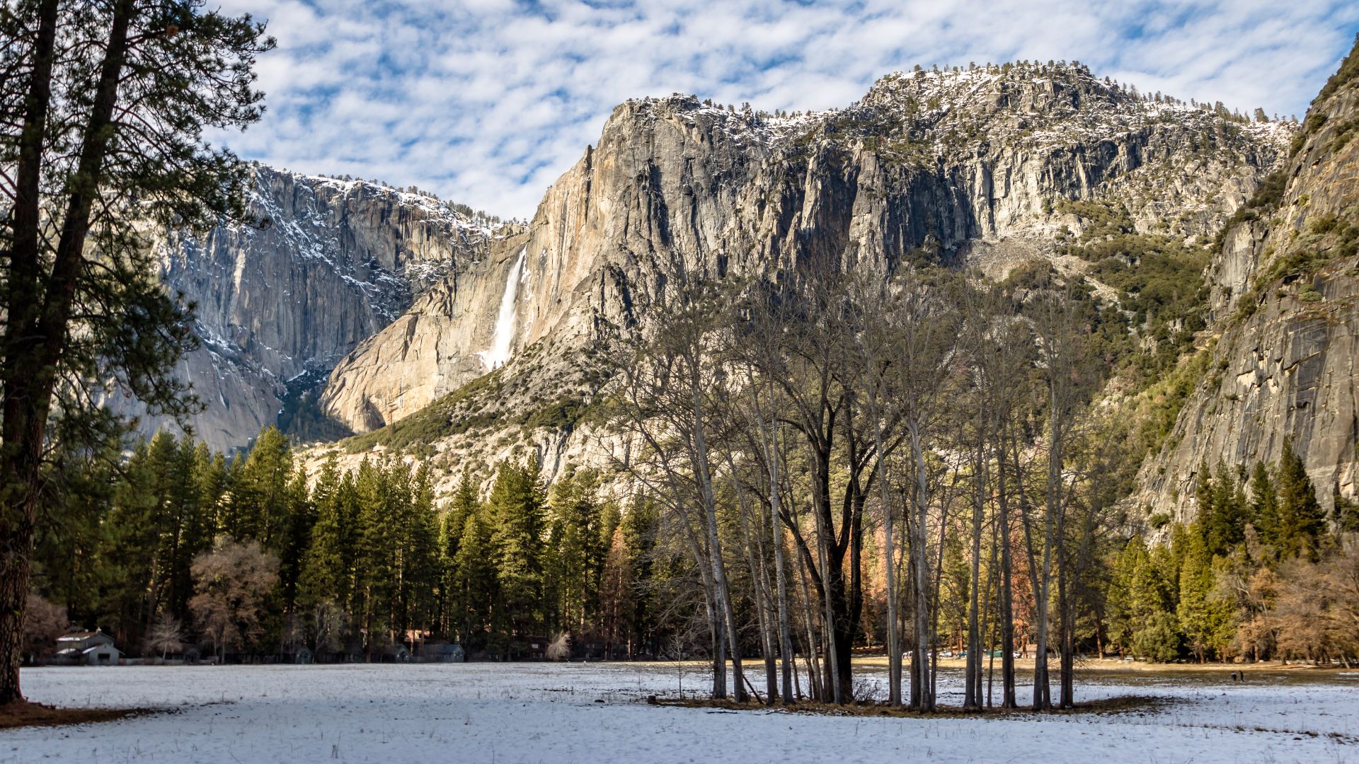 A wide shot of Yosemite Falls cascading down a massive granite cliff face, surrounded by snow-dusted ground and tall evergreen trees, under a partly cloudy sky.