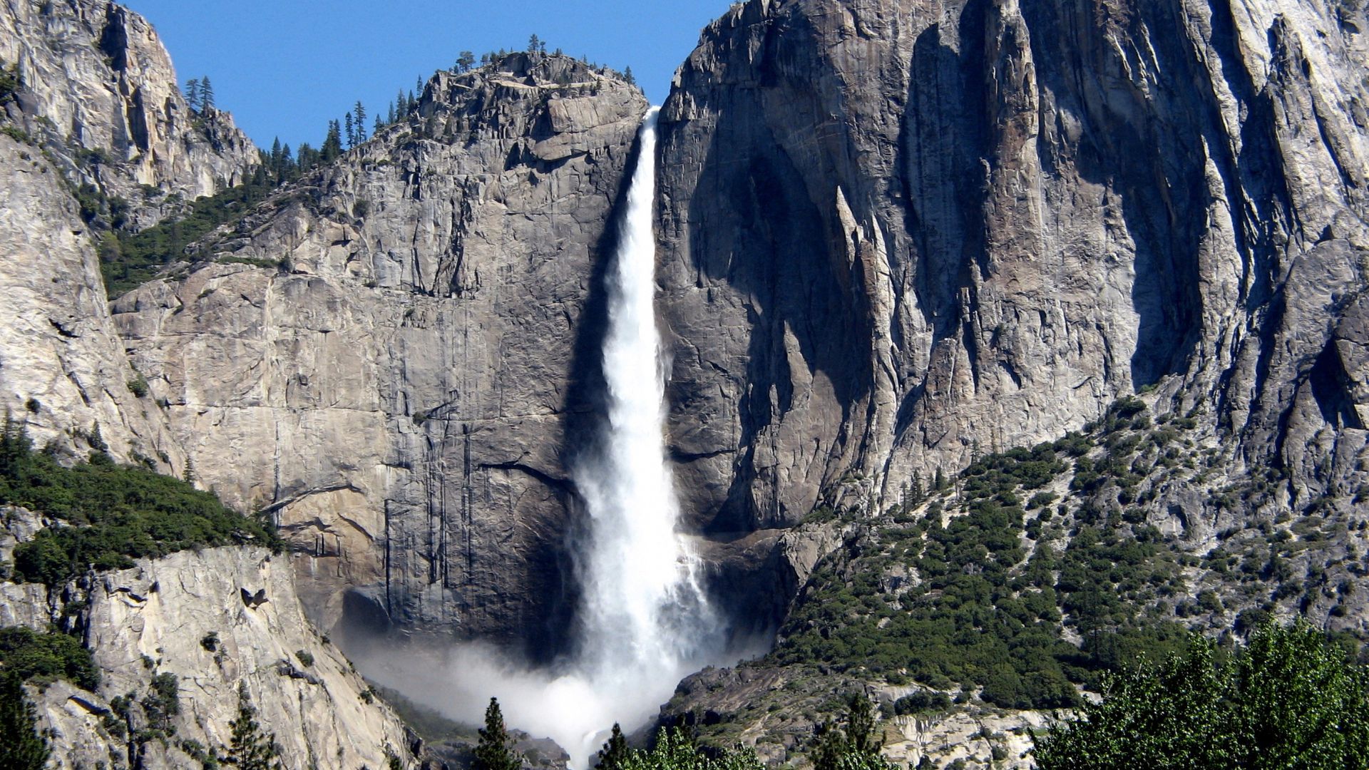 A majestic waterfall cascades down a massive, rugged granite cliff face, surrounded by lush green trees at its base and on the lower slopes of the mountains in Yosemite National Park. The sky above is clear and blue.