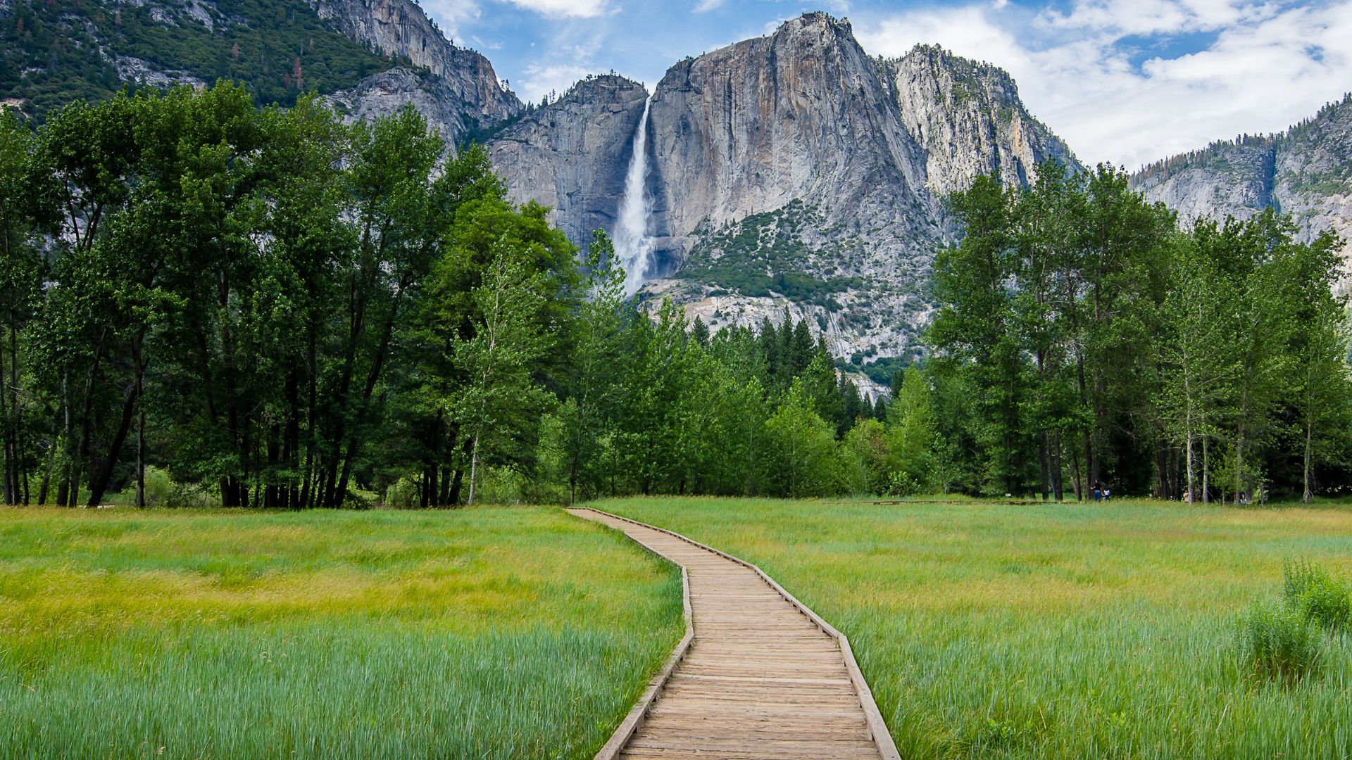 A wooden boardwalk path winds through a vibrant green meadow towards a towering granite cliff face with a prominent waterfall cascading down in the distance, framed by lush green trees on either side under a partly cloudy sky.