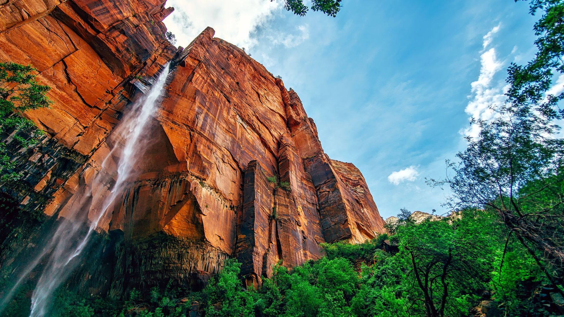 A powerful waterfall cascades down a massive, reddish-brown rock face under a bright blue sky with scattered clouds, surrounded by lush green trees at the base.