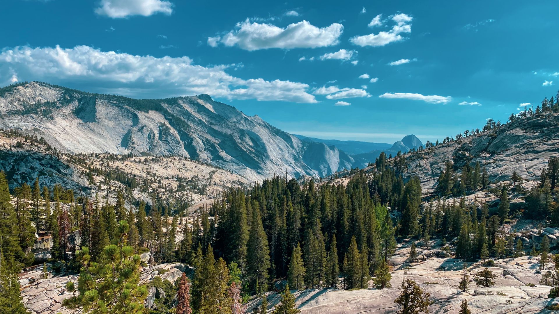 A wide-angle landscape shot of Yosemite National Park, featuring towering granite mountains under a blue sky with scattered clouds, dense evergreen forests in the midground, and rocky terrain in the foreground.