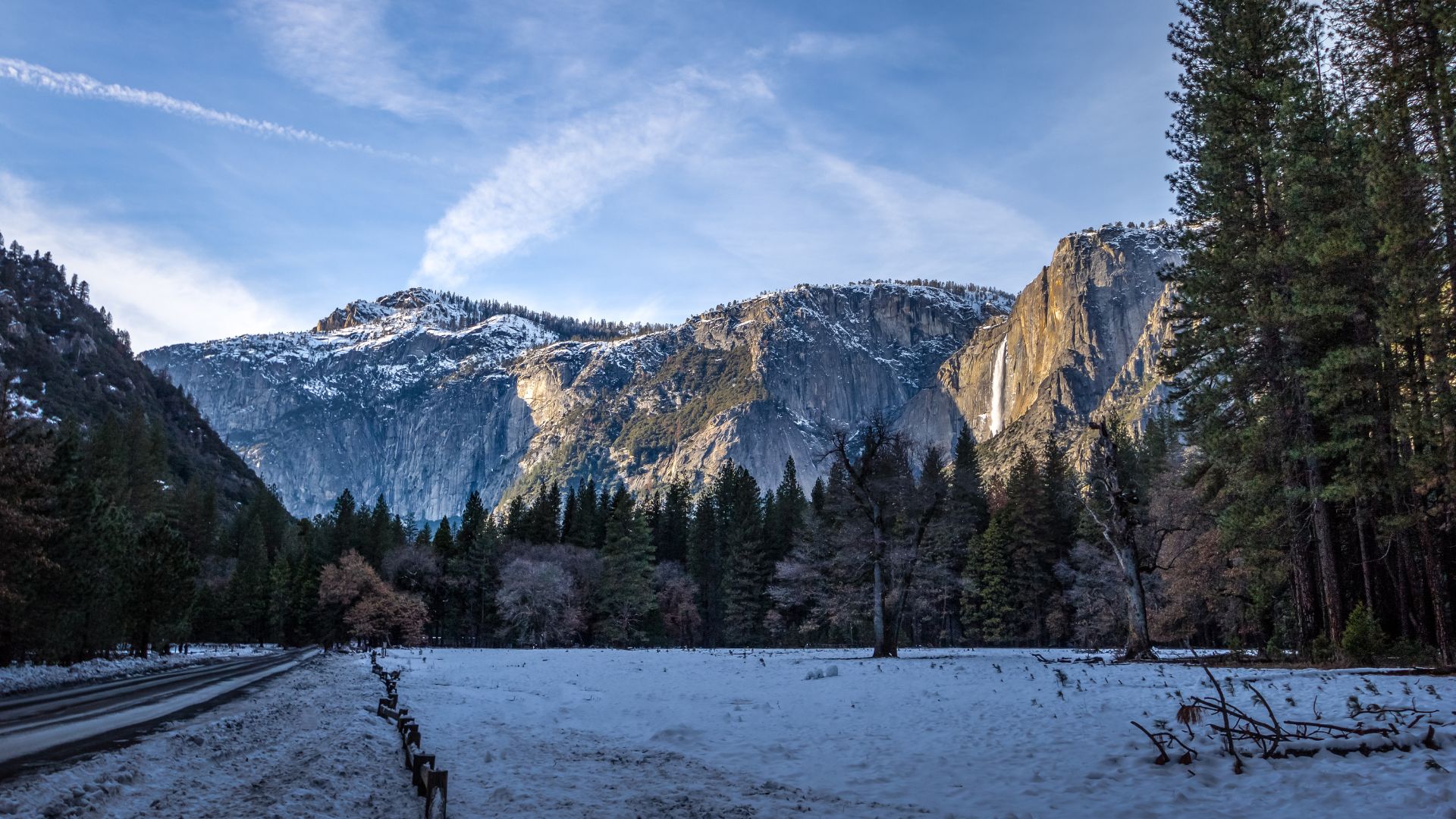 A wide shot of a snow-covered Yosemite Valley in winter, with towering granite cliffs and snow-capped mountains in the background, evergreen trees lining the valley floor, and a visible waterfall (likely Yosemite Falls) cascading down a cliff face on the right, under a clear sky with faint contrails.