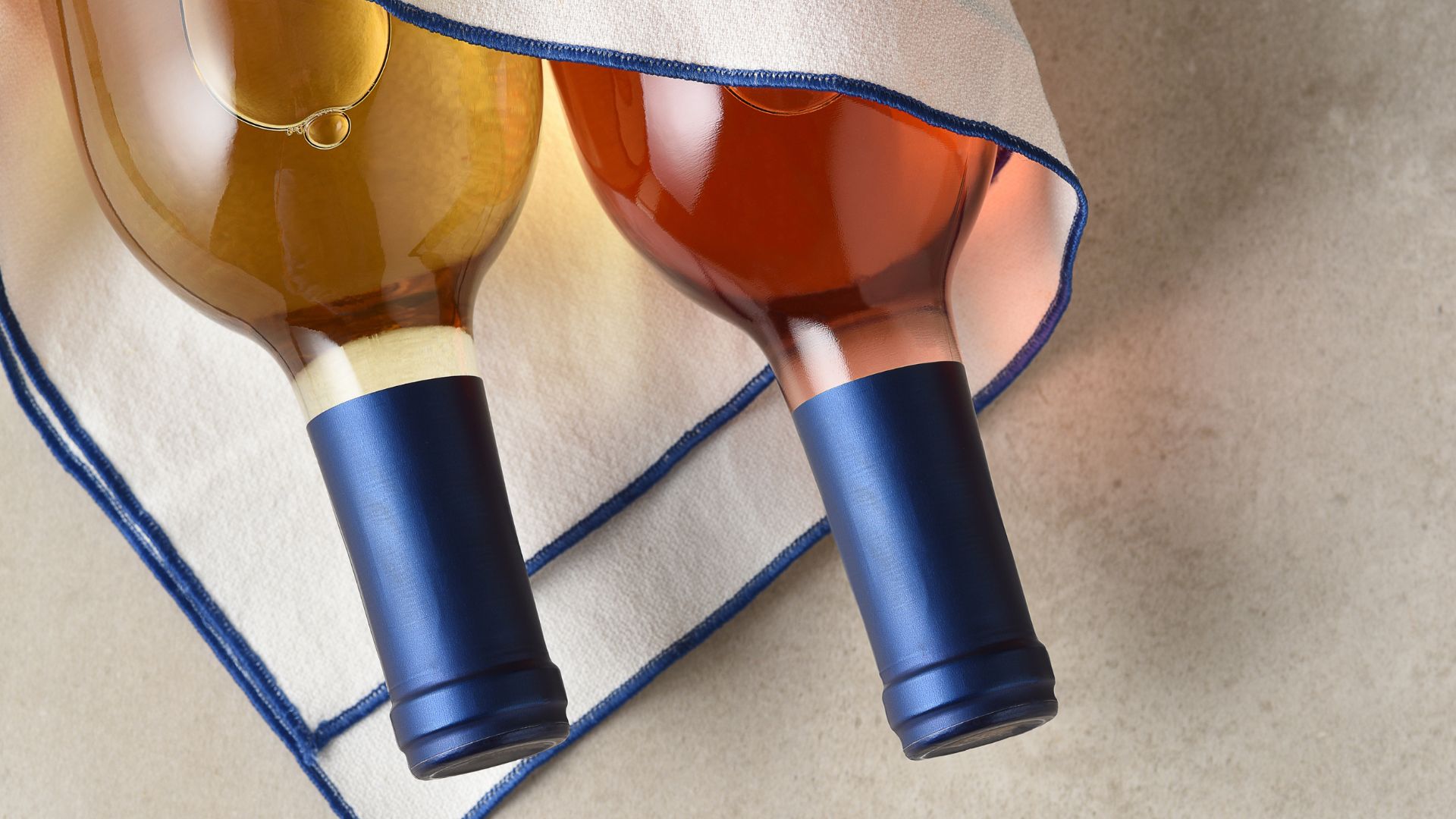 A close-up overhead shot of two wine bottles, one containing golden-hued white wine and the other a reddish-amber rosé or red wine, both featuring dark blue foil capsules, resting on a folded white napkin with a blue embroidered edge, against a light-colored textured background.